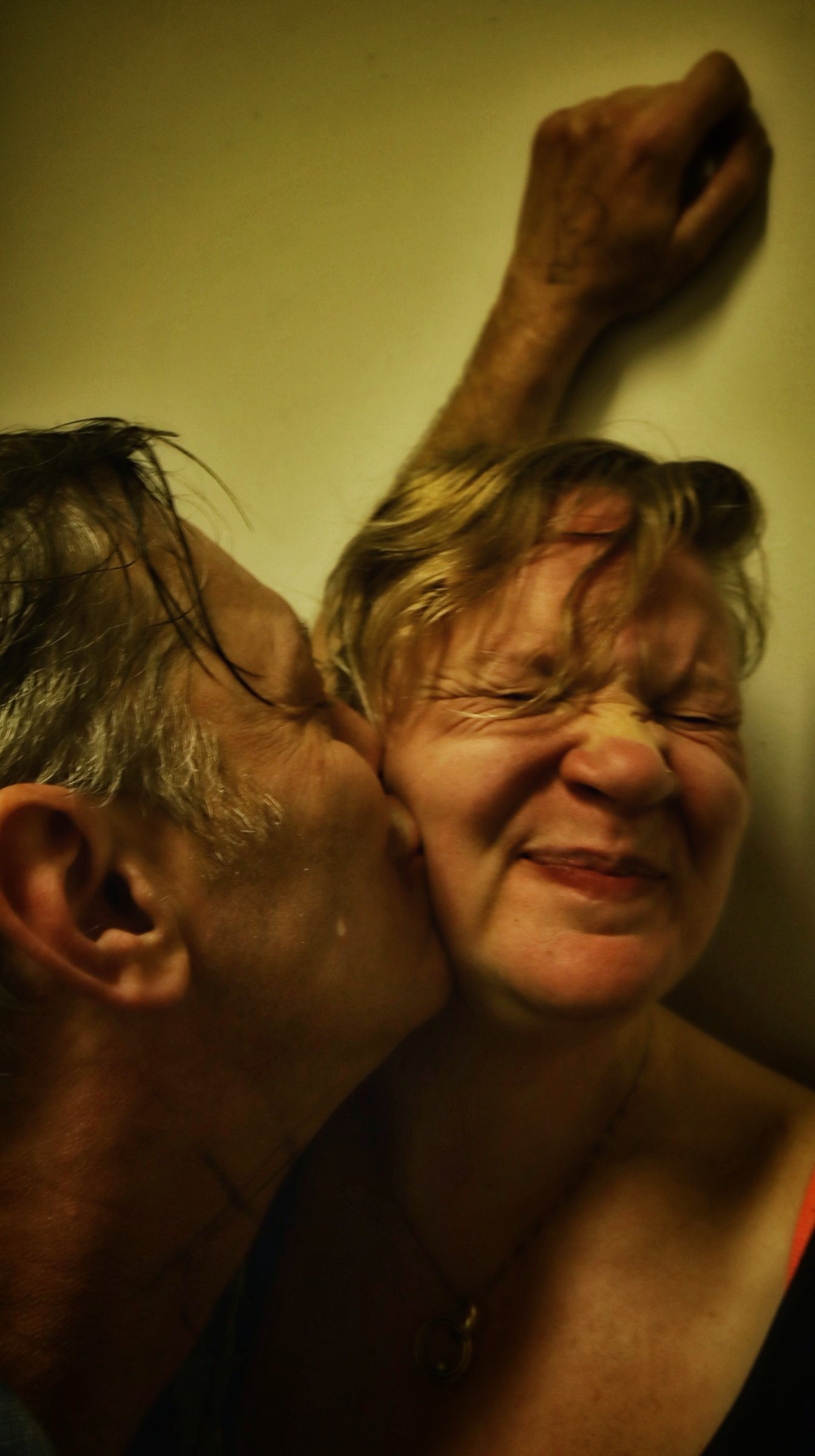 A man is kissing a woman on the cheek while she smiles with her eyes closed. The woman has short hair, and the man has glasses and short hair. There is a wall with a wooden sculpture of a hand in the background.
