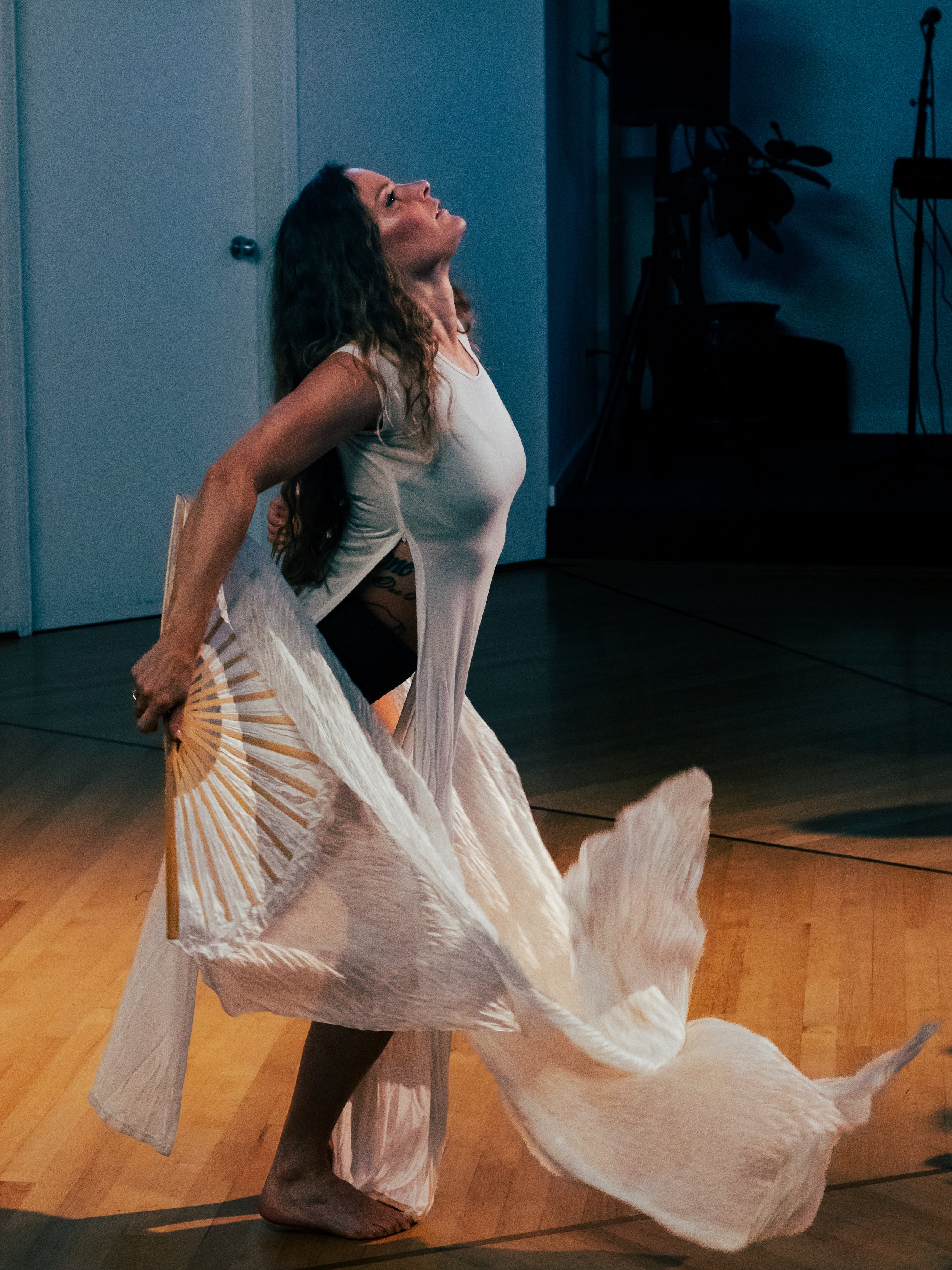 A woman with curly hair performing a dance in a studio, wearing a light-colored flowing dress, with a focused expression, and a stage setup with a microphone and potted plant in the background.
