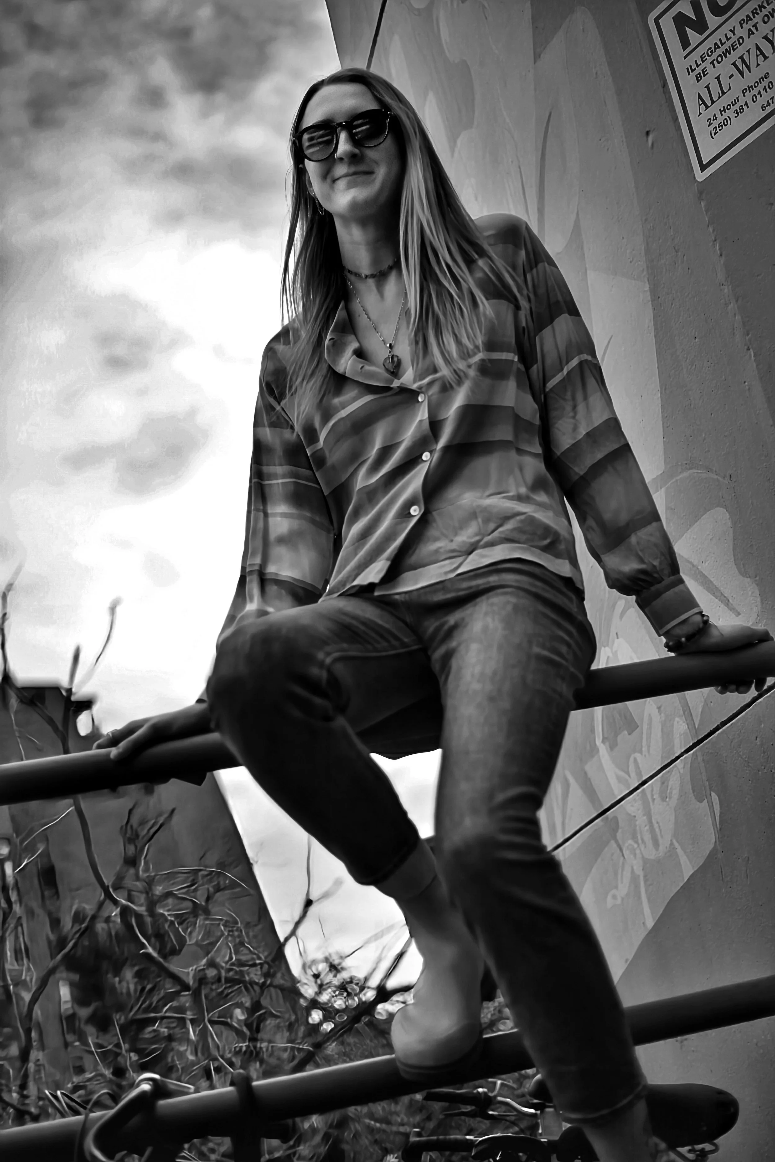 A woman wearing sunglasses and jewelry, dressed in a checked shirt and jeans, sitting on a railing outdoors against a wall with a sign, with a cloudy sky in the background.