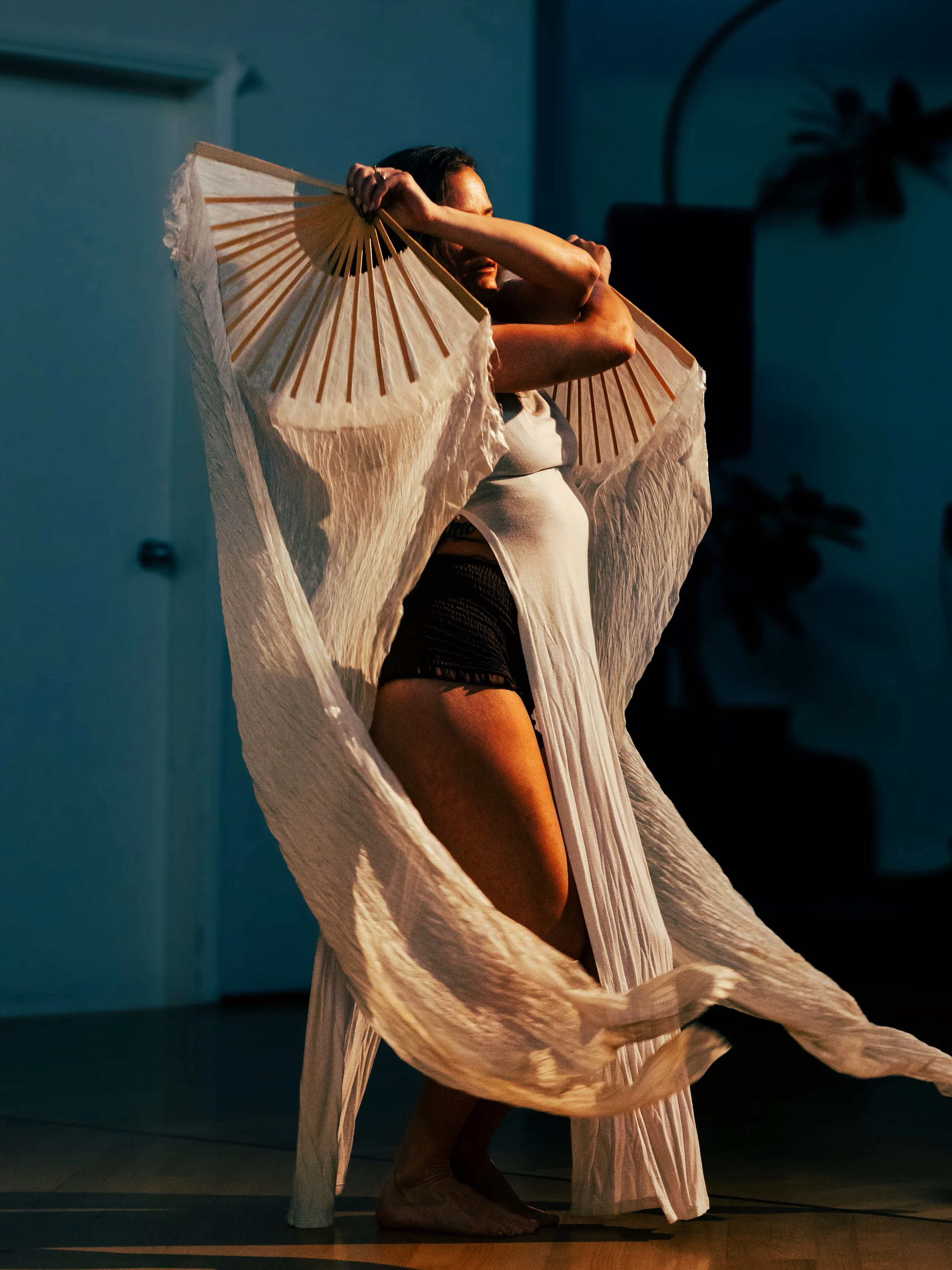 Woman dancing with a large white flowing fabric and reaching over her head, set against a dark background.