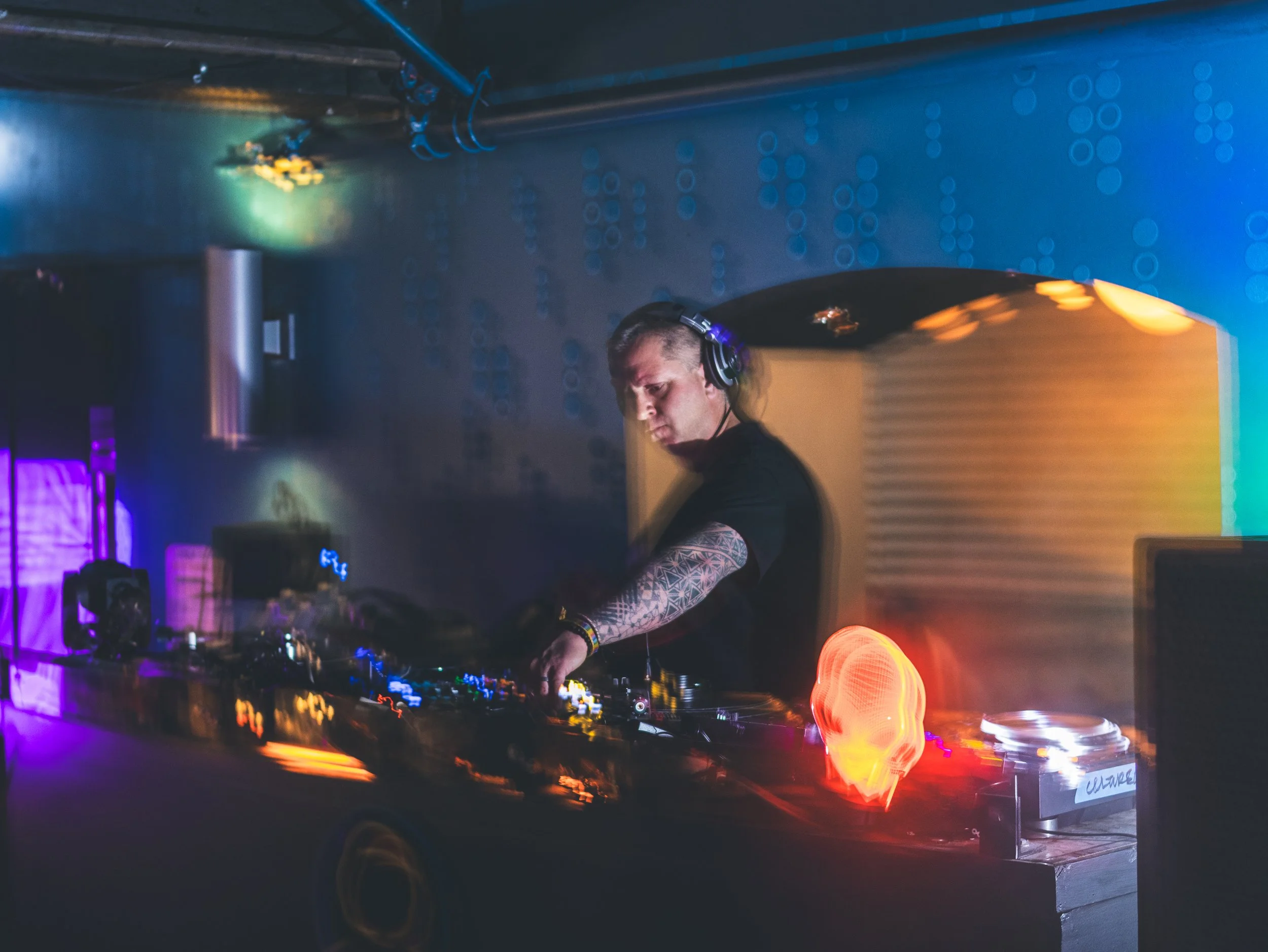 A DJ with long hair and tattoos on his arm performing at a nightclub, with colorful lights and blurred motion effects.