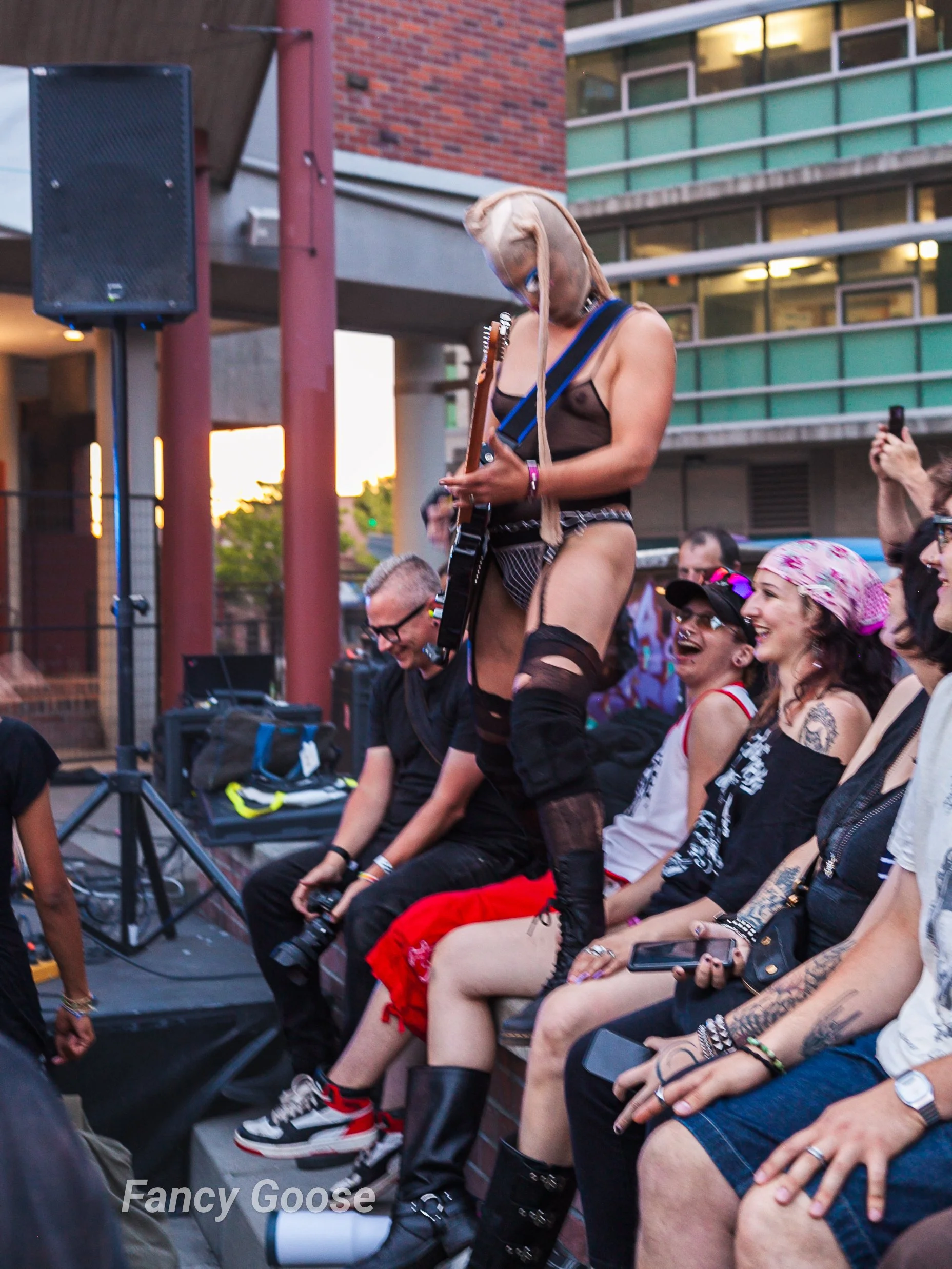 A female guitarist with long dreadlocks and blue eye makeup performing on stage at an outdoor event, surrounded by audience members, at sunset in an urban setting.