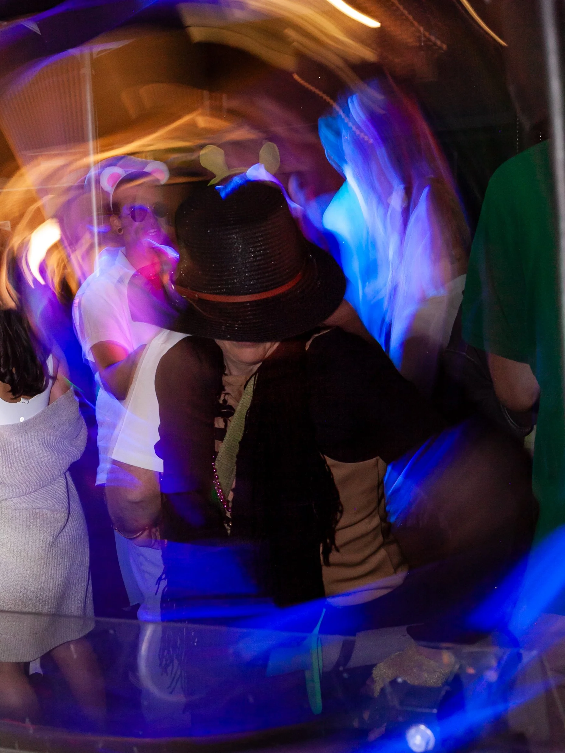 People in costumes, including one with bunny ears and a black hat, at a party or celebration with colorful lights and motion blur.