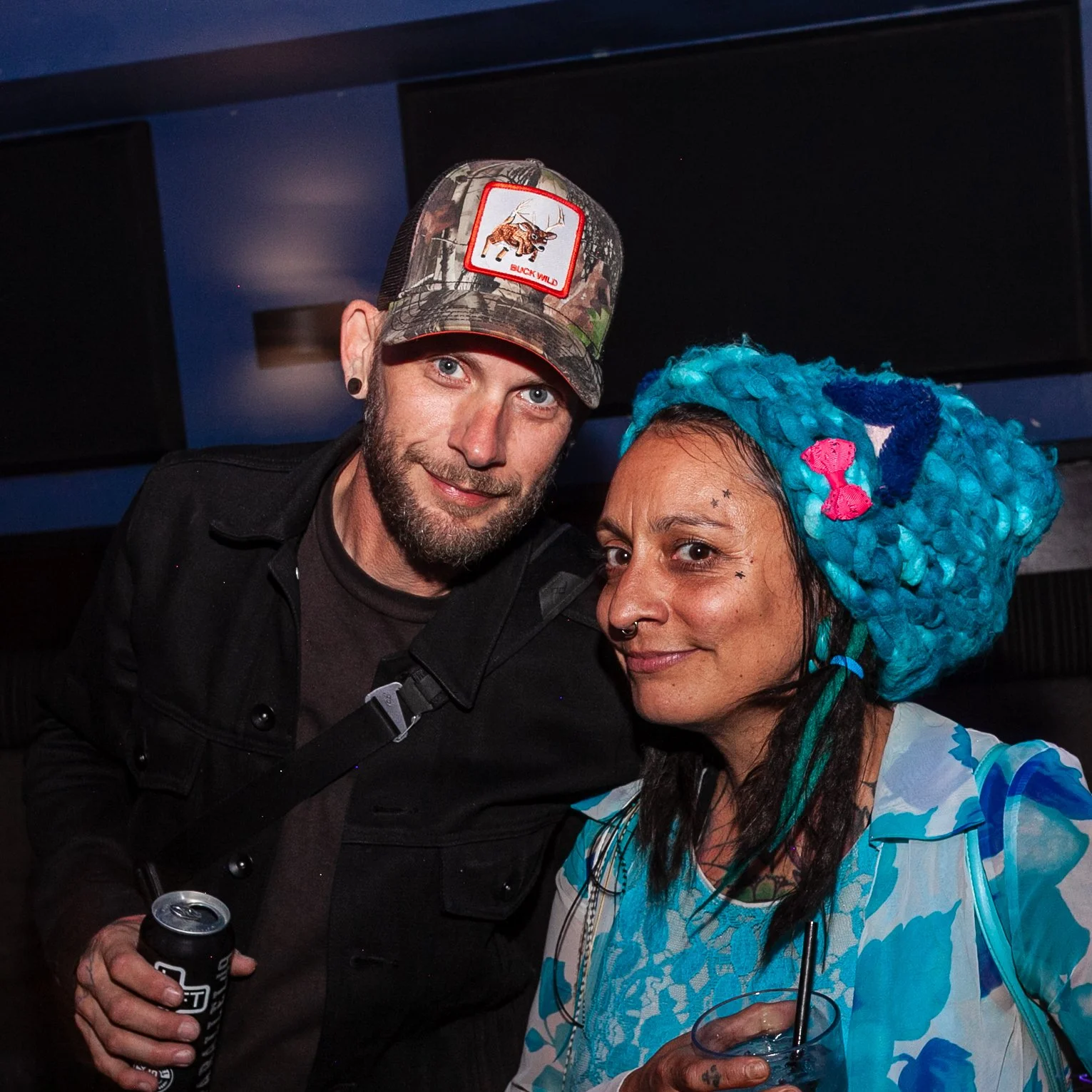 Two people smiling and posing close together, one man wearing a camouflage baseball cap with a wild animal patch and a woman with bright blue dreadlocks, holding drinks and standing against a dark background.