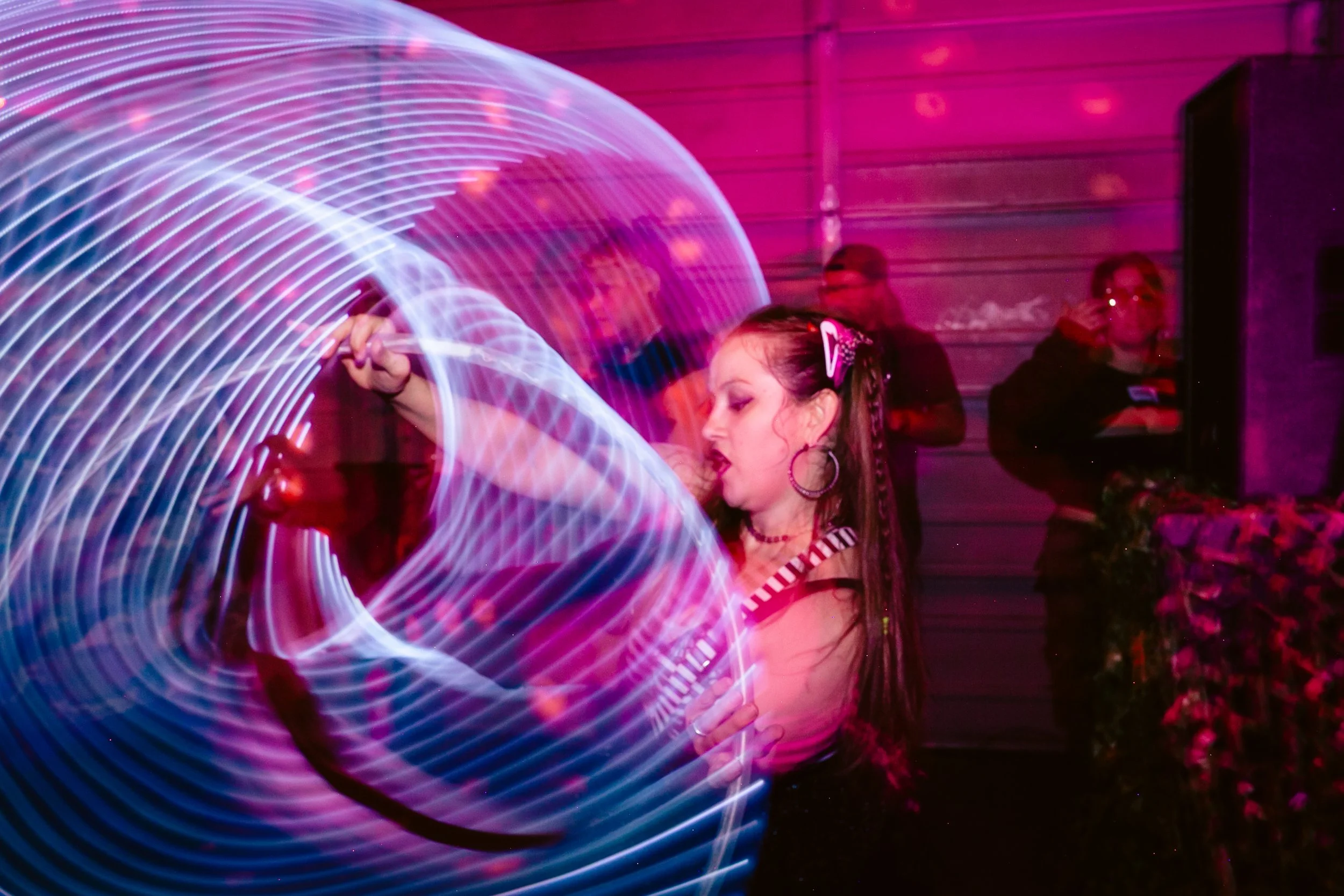 A young girl with braided hair performing with light trails creating circular patterns in a dimly lit indoor setting, with a woman and a man observing in the background.