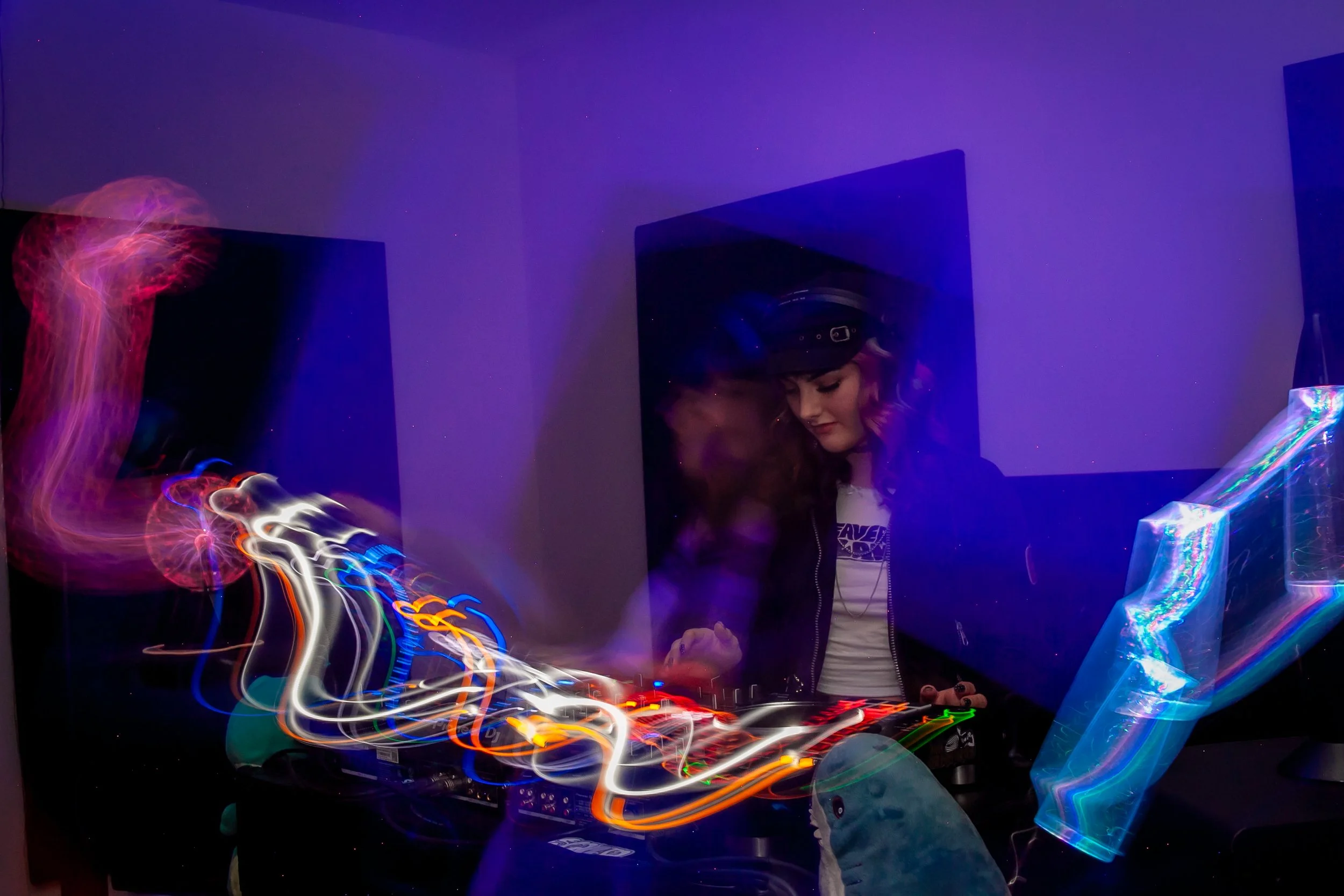 Person with curly hair wearing headphones, standing behind DJ equipment with colorful light trails in a dark room with purple walls.
