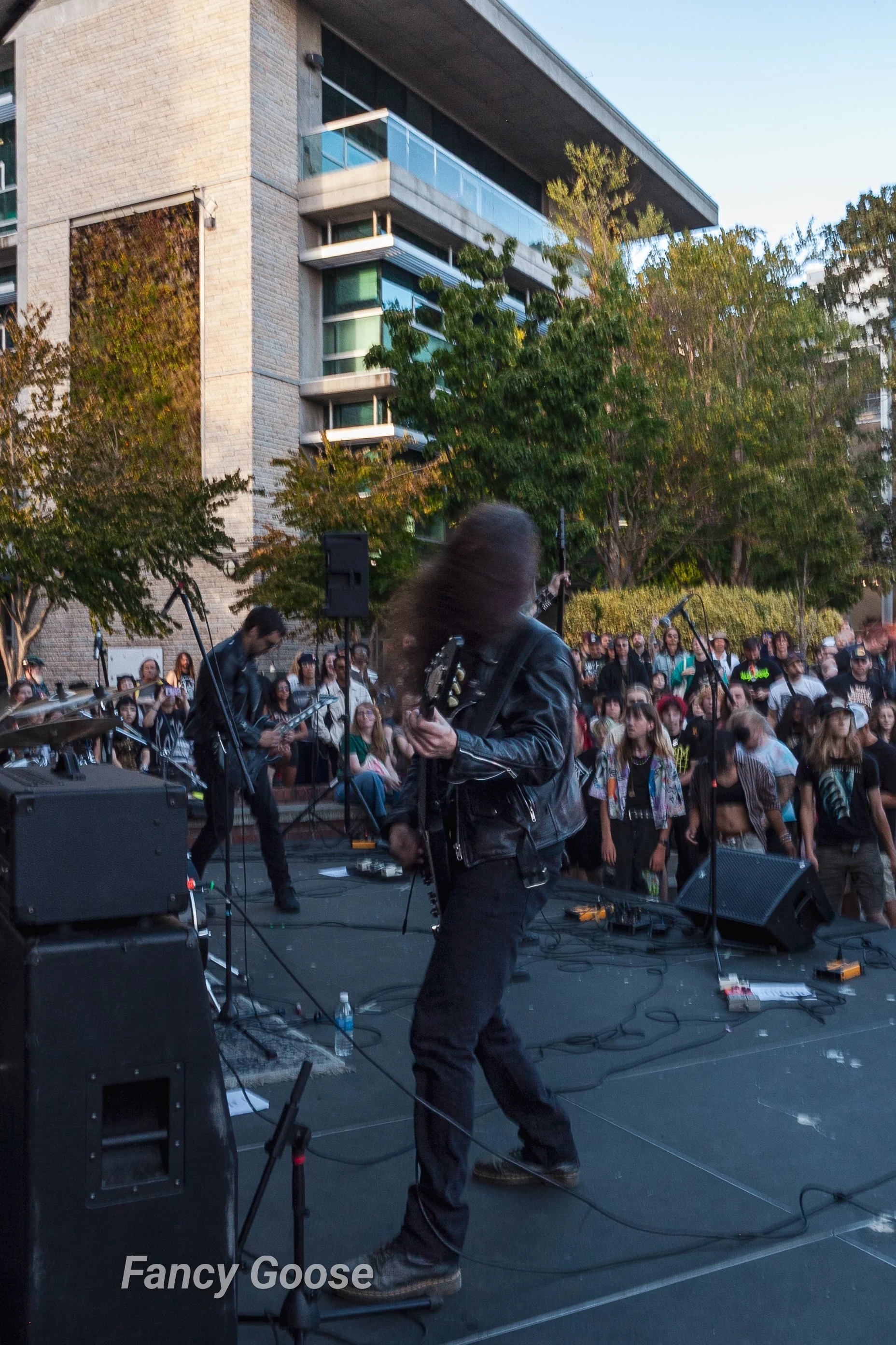 A street performance with musicians playing instruments on a stage, crowd watching, in an urban area with tall apartment buildings and trees, during the daytime.