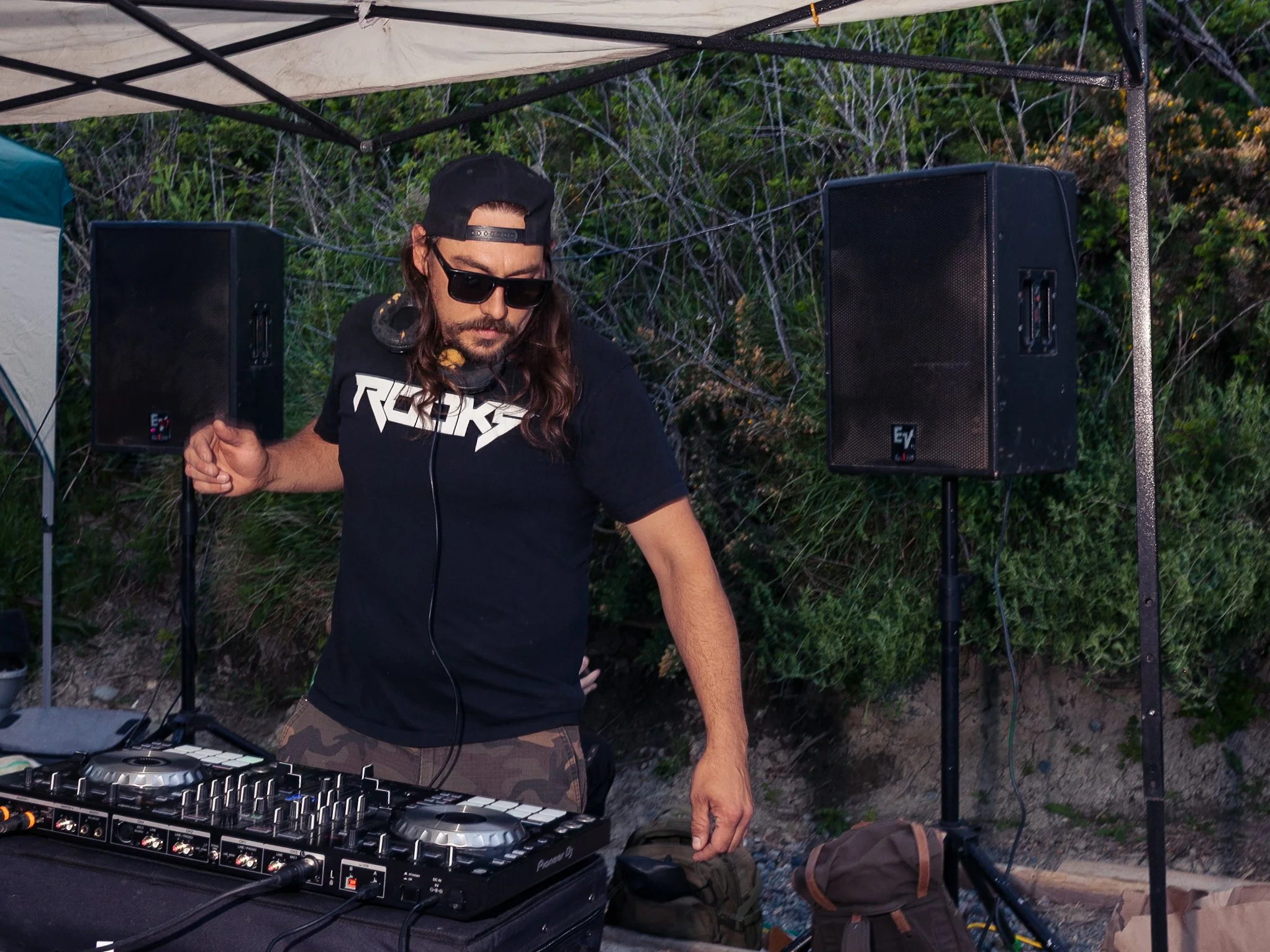 A DJ with long hair, sunglasses, and a black T-shirt with a logo is performing outdoors at night under a canopy, surrounded by speakers and electronic equipment.