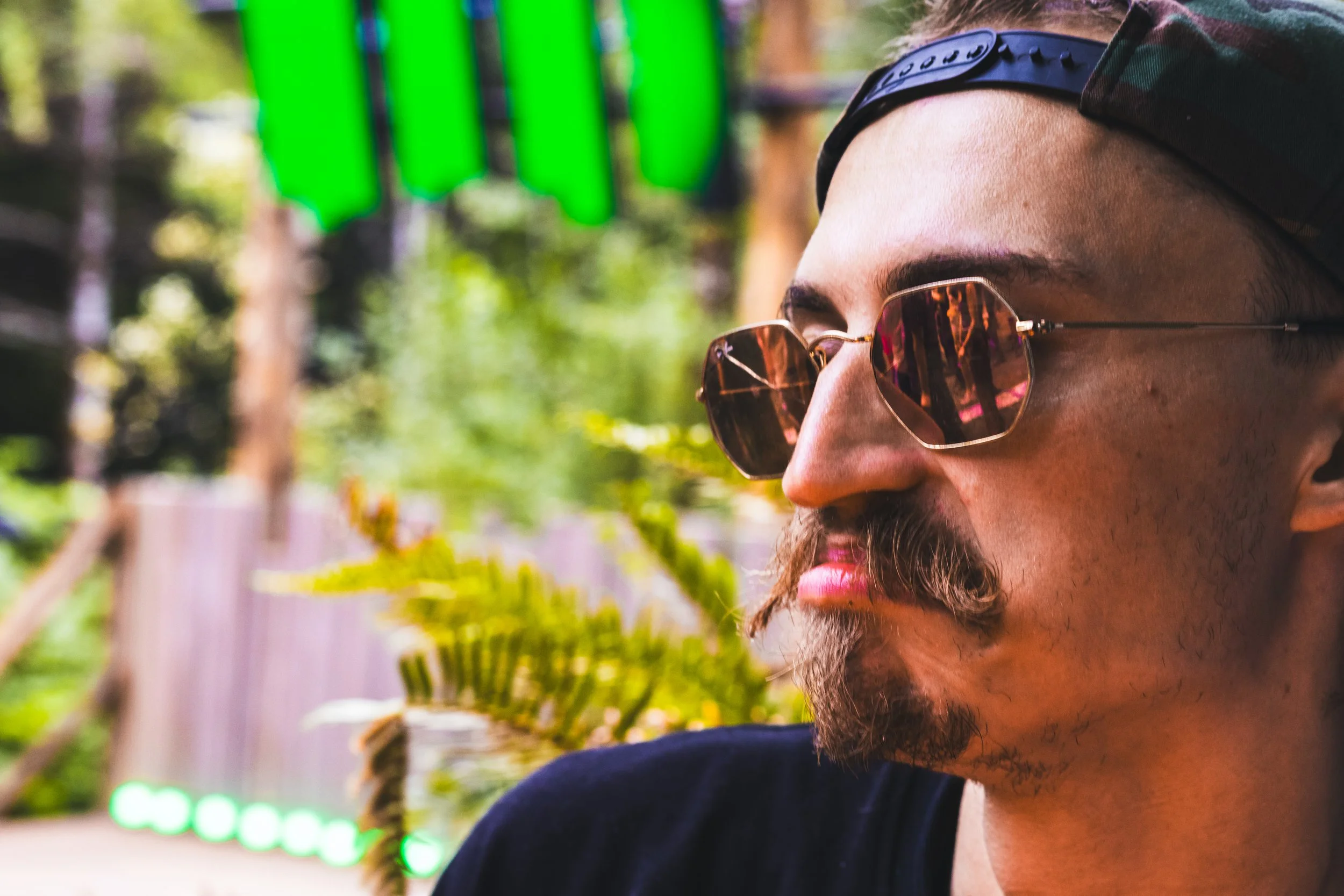 A close-up of a man with a beard wearing sunglasses and a camouflage cap, standing outdoors in a park or wooded area, with green string lights visible in the background.