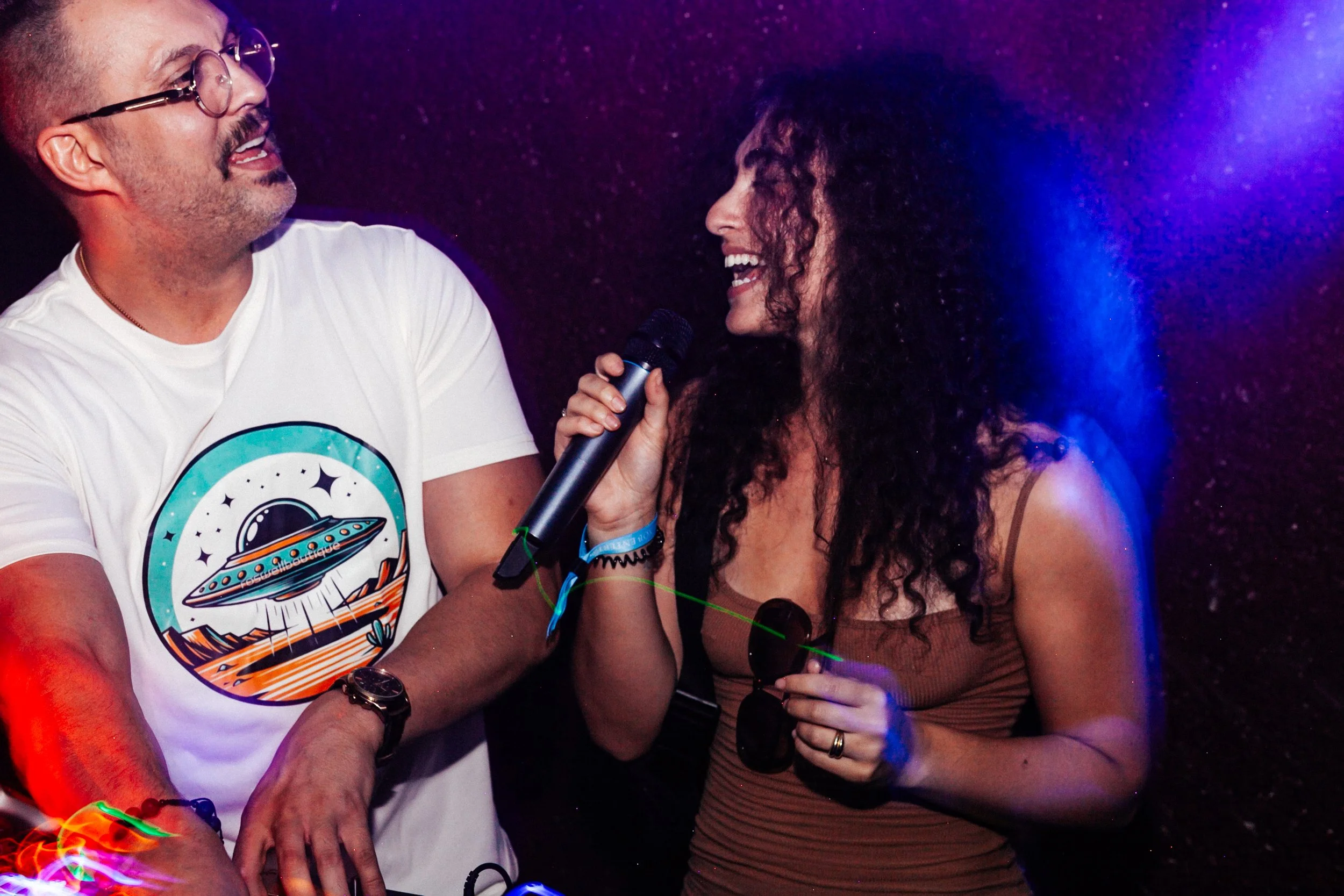 A man and woman are happily singing into a microphone together. The man is wearing glasses and a white t-shirt with a space-themed design. The woman has curly hair and is smiling brightly. They are in a dark room with colorful lights.