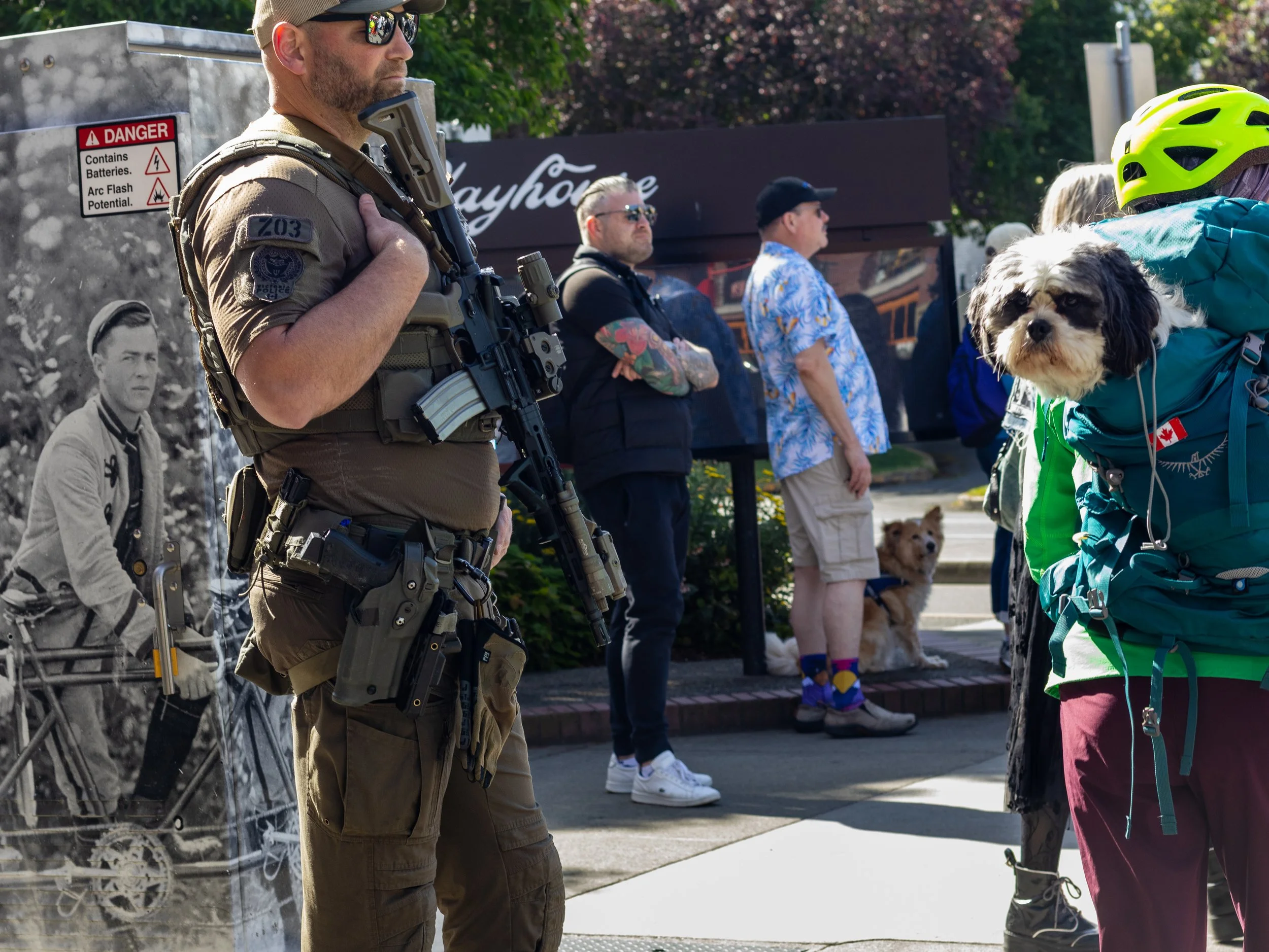 A man in tactical gear and sunglasses holding a rifle stands on a street, with a group of people, some with dogs, standing in the background.