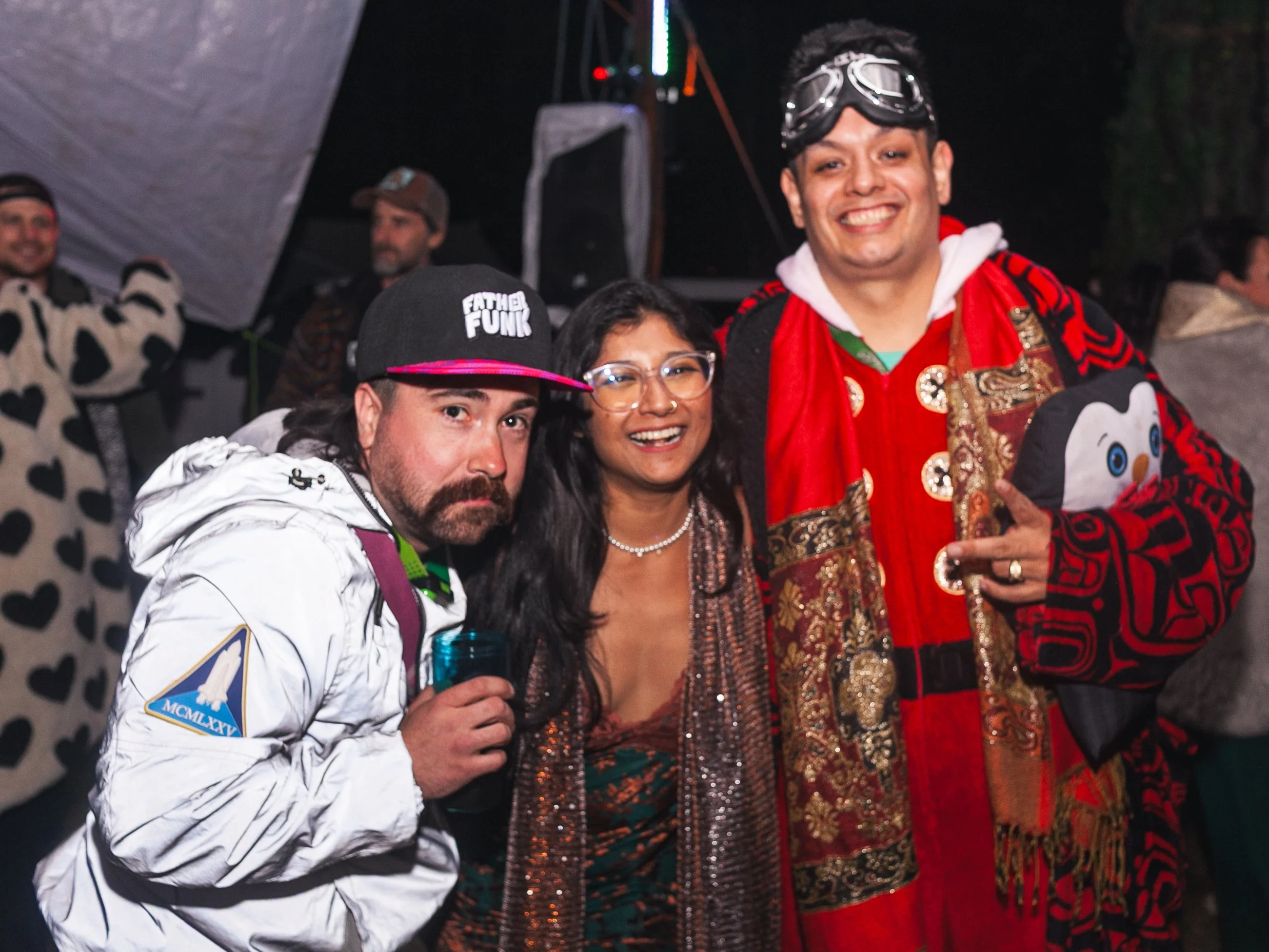 Group of people at an outdoor event at night, including a man in a white jacket with an NASA patch, a woman with glasses and a nose ring, and a man in a colorful, patterned red and gold jacket with goggles on his head, smiling.