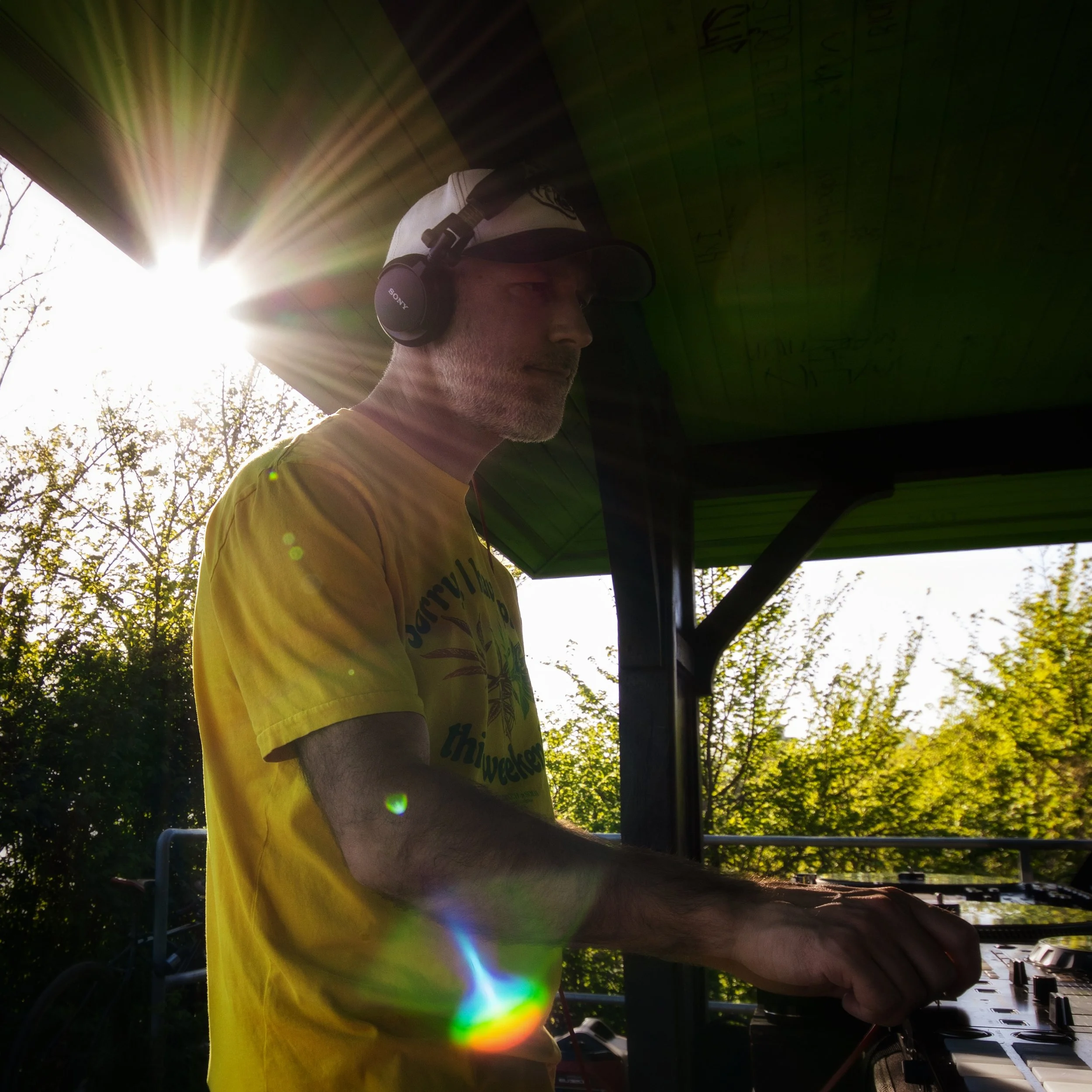 A man wearing headphones and a yellow T-shirt is outdoors working with electronic equipment. The sun shines brightly, creating lens flare and casting light through the trees in the background.