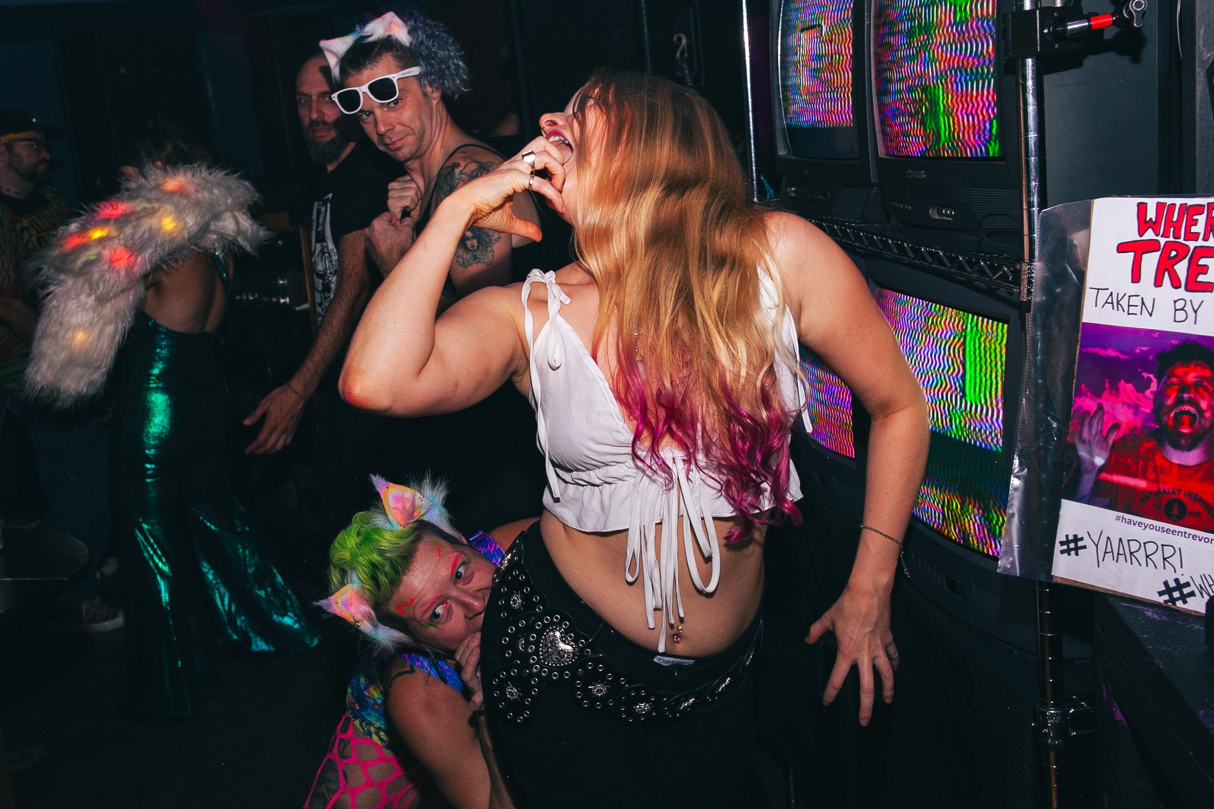 Group of people partying at a club, dressed in colorful and eccentric outfits, with neon and laser lighting effects.