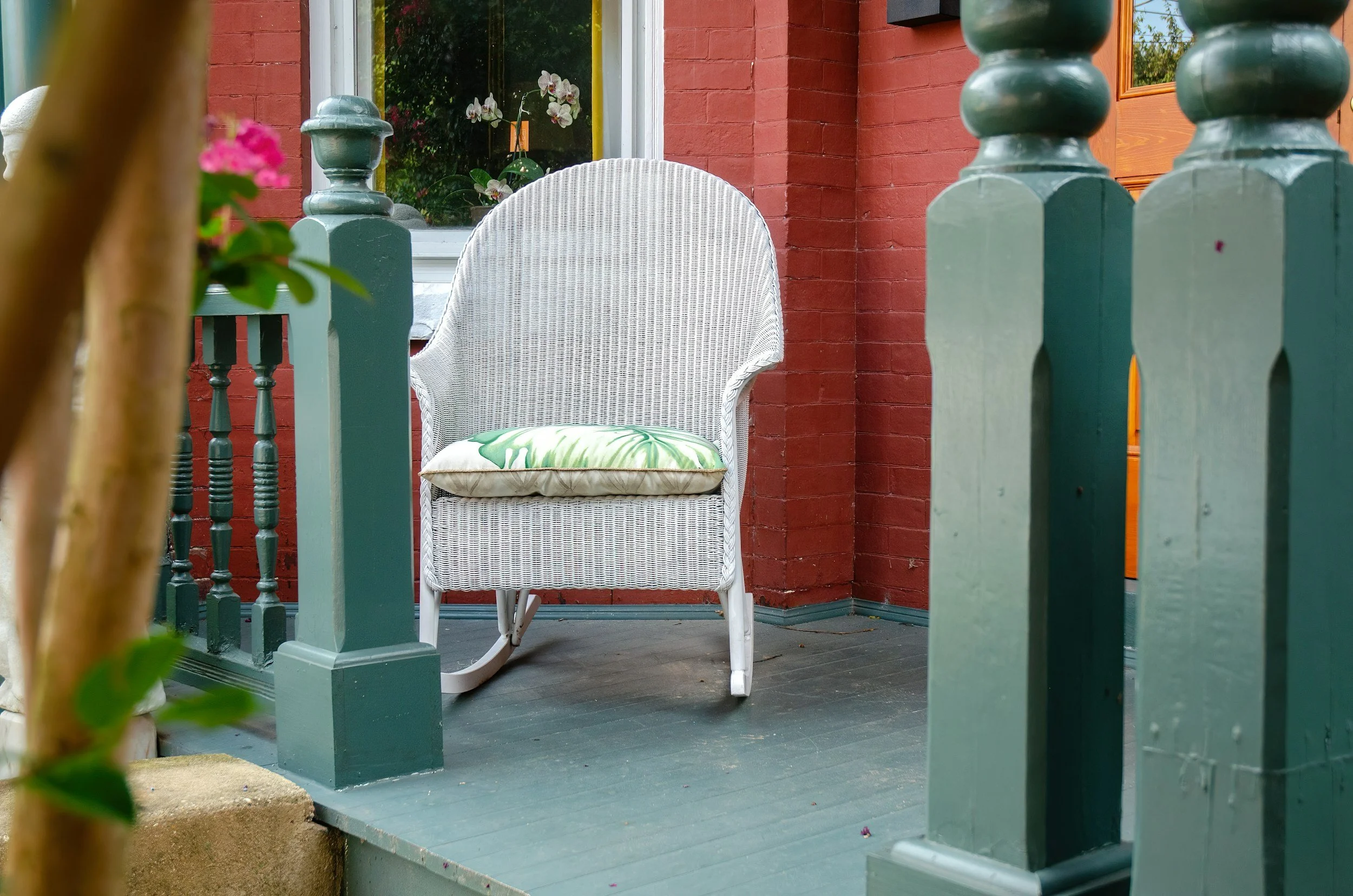 Front porch of a house painted red with a white wicker chair on the painted teal porch floor. The porch is decorated with teal picket fence-style railing and a small potted plant with pink flowers in the foreground. A window with orchids inside is visible on the house wall.