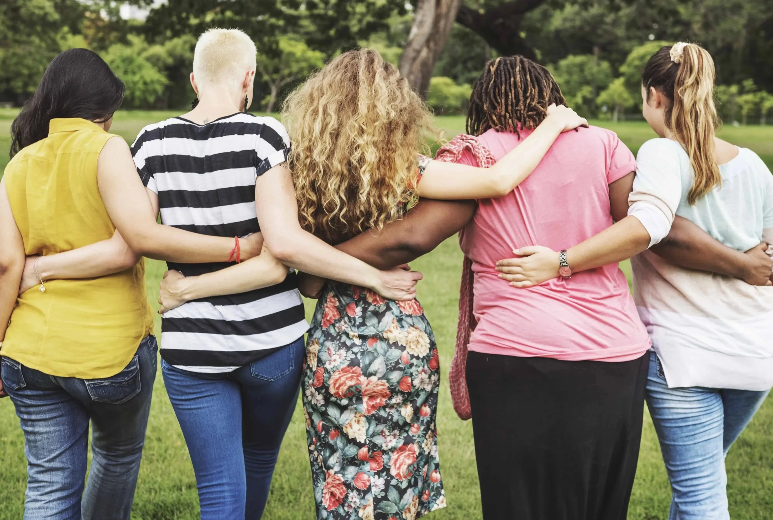 Group of diverse women standing closely together outdoors on green grass, with arms around each other's shoulders, facing away from the camera in a park.