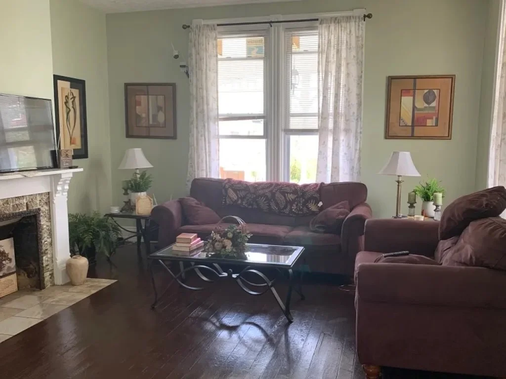 Living room with light green walls, large windows with white curtains, a dark sofa, an armchair, a coffee table with books and flowers, a fireplace with a vase nearby, and wall art decorations.