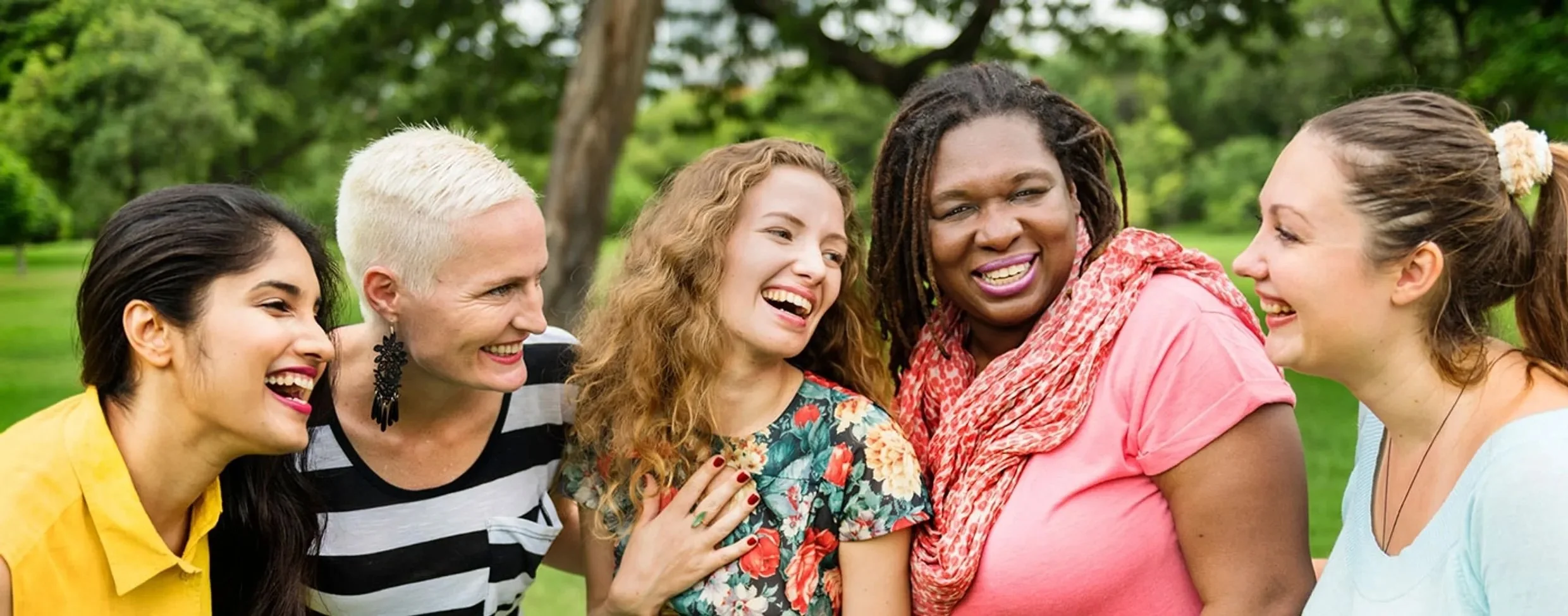 Group of five women laughing and talking outdoors in a park with green trees in the background.