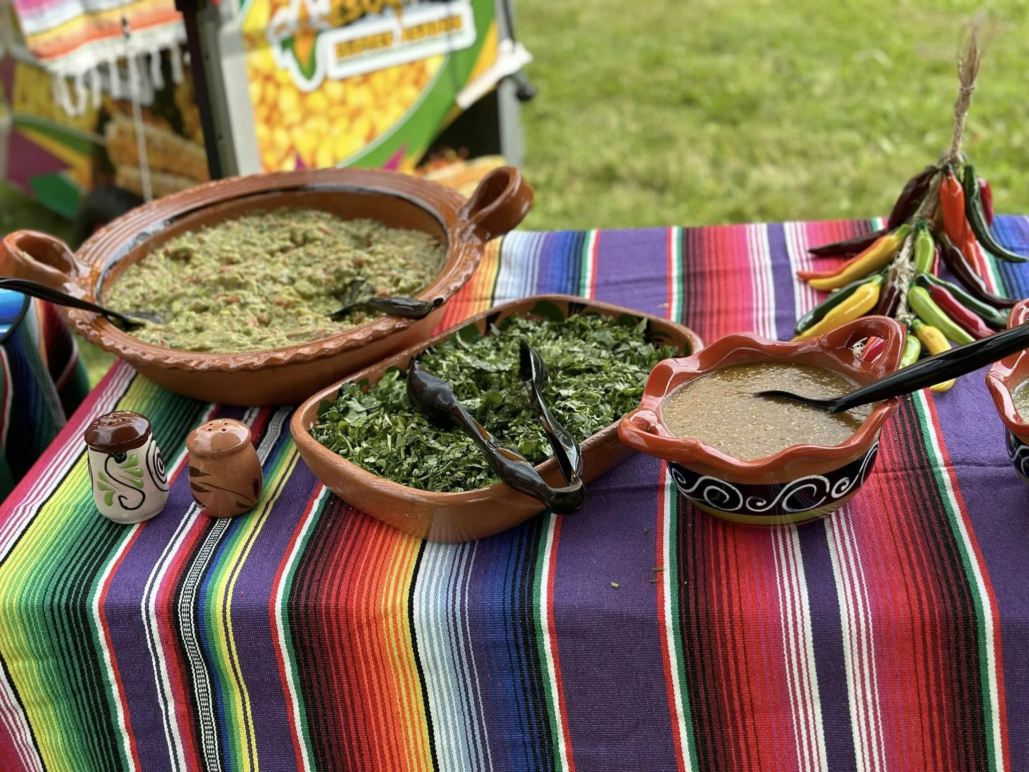 A colorful outdoor Mexican food display with bowls of guacamole, salsa, and chopped herbs on a vibrant striped tablecloth, with chili peppers and decorative items.