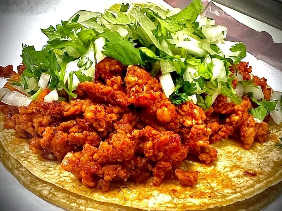 Taco with seasoned ground chicken, shredded lettuce, and chopped onions on a yellow corn tortilla.