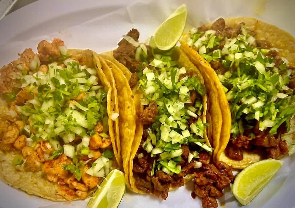 Three tacos filled with seasoned meat and topped with chopped onions and cilantro, garnished with lime wedges.
