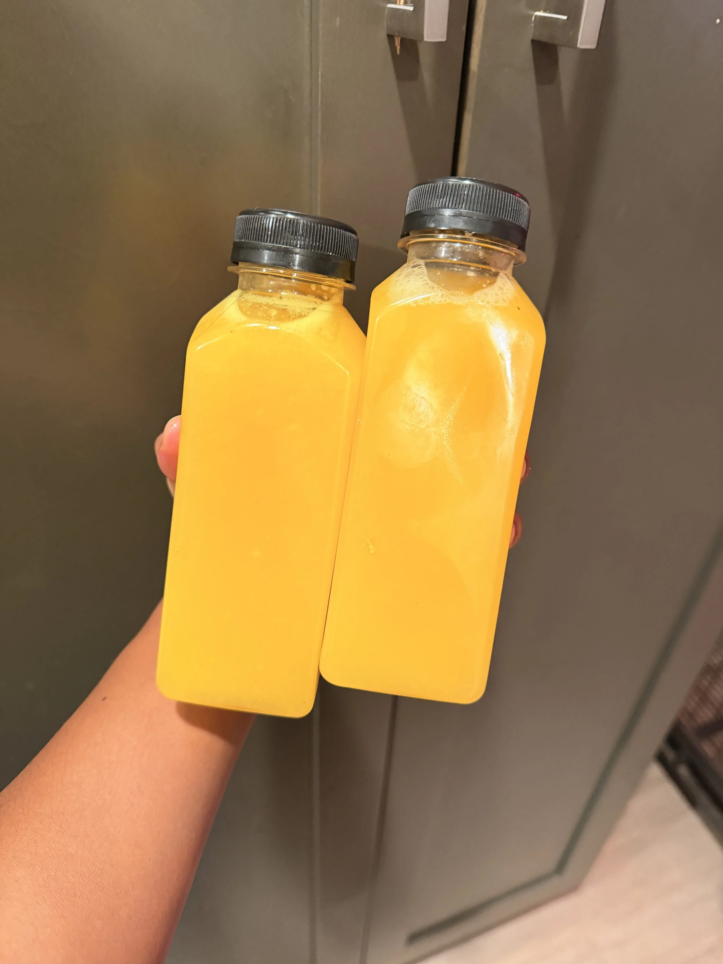 Two small bottles of orange juice with black caps are being held up in front of a gray-colored metal locker or cabinet.