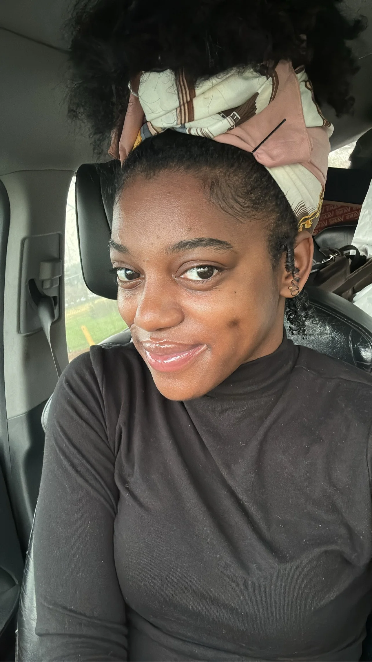 A woman sitting in a vehicle, taking a selfie. She has a smile, wearing a black top, with curly hair pulled back, and is seated near the window with a view of some greenery outside.