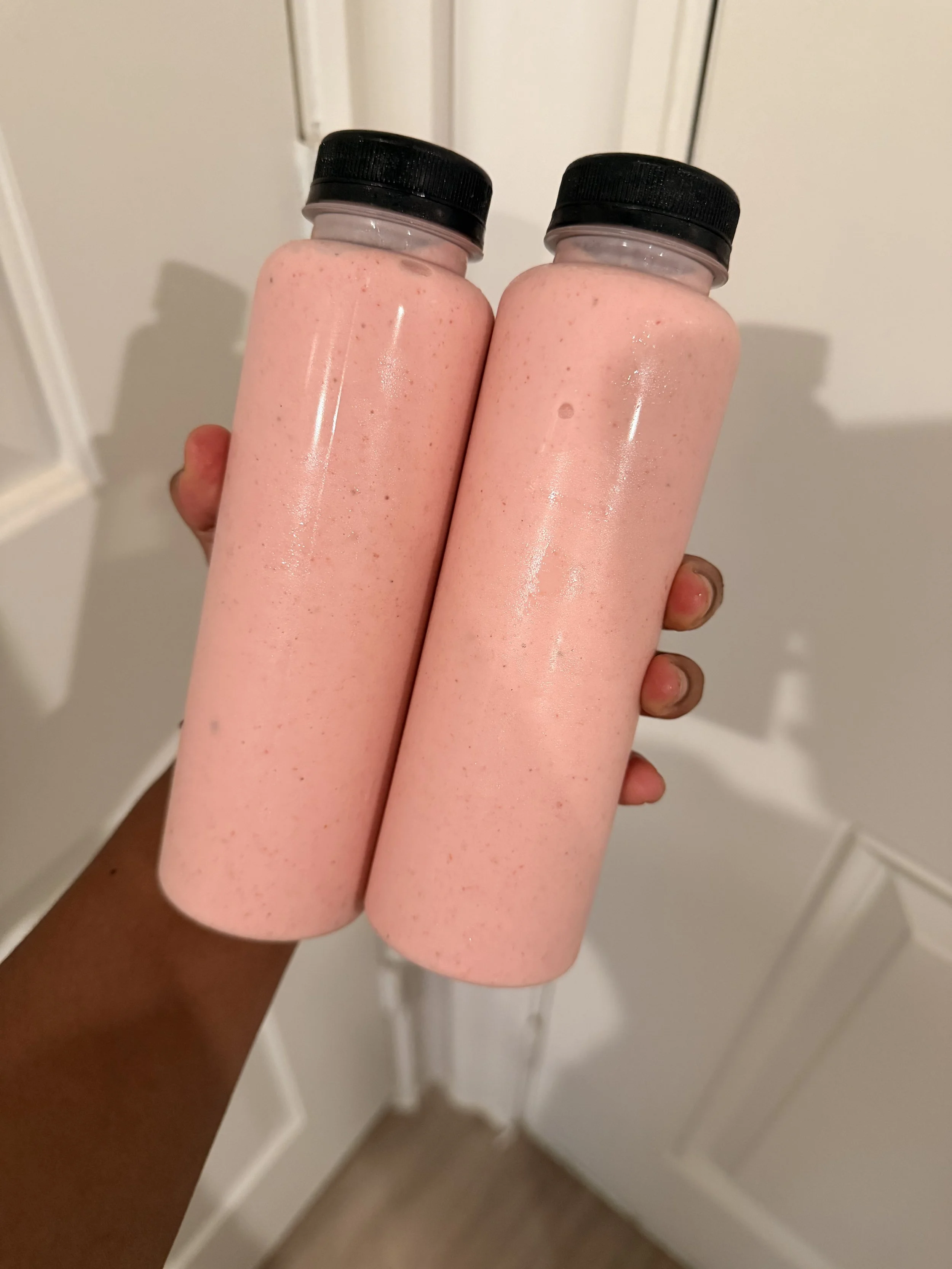 Two bottles filled with pink smoothie or milkshake being held up in front of a white door.