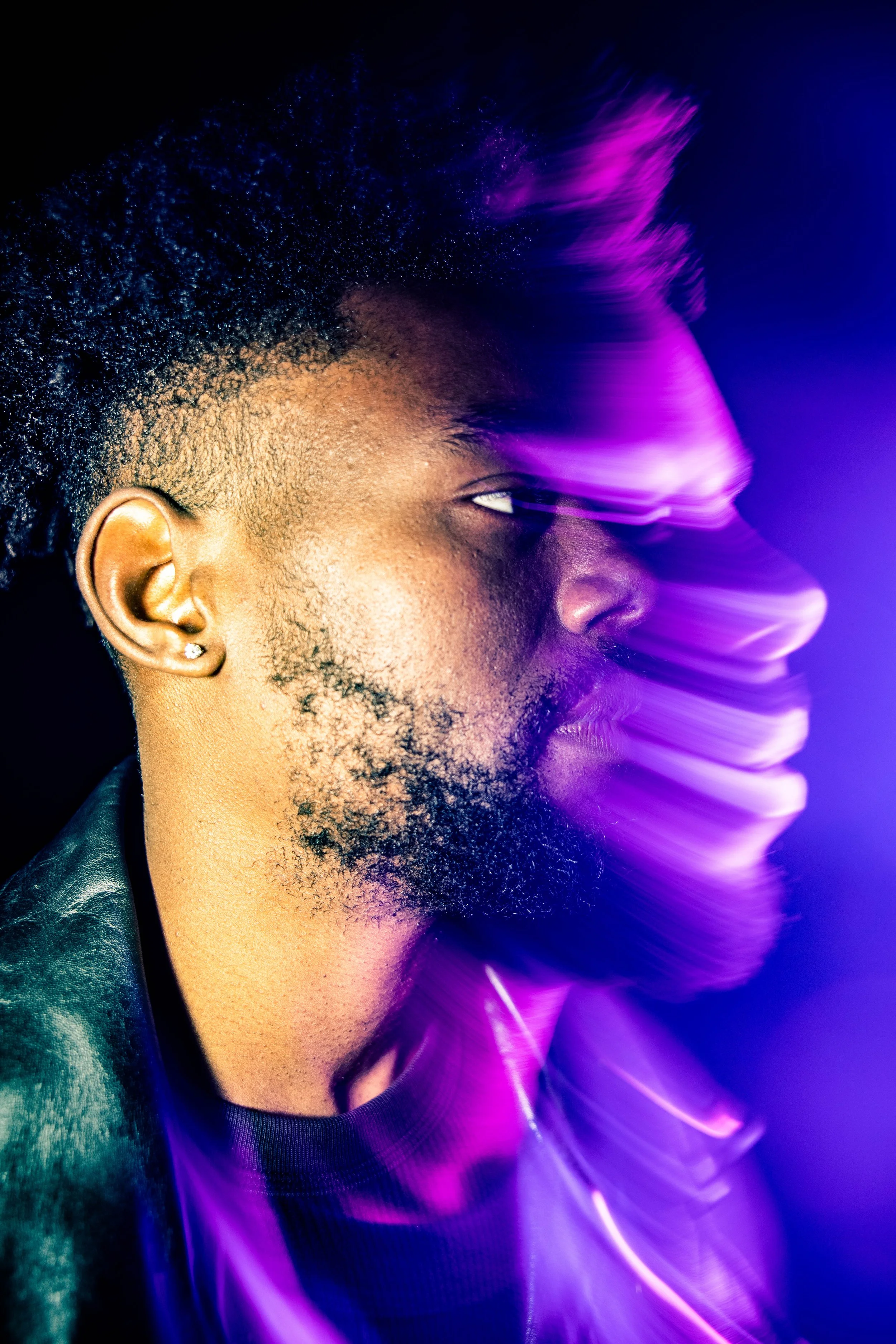 Close-up of a man's face in profile with dark skin, textured hair, and a beard and earrings, illuminated by purple and blue light effects.