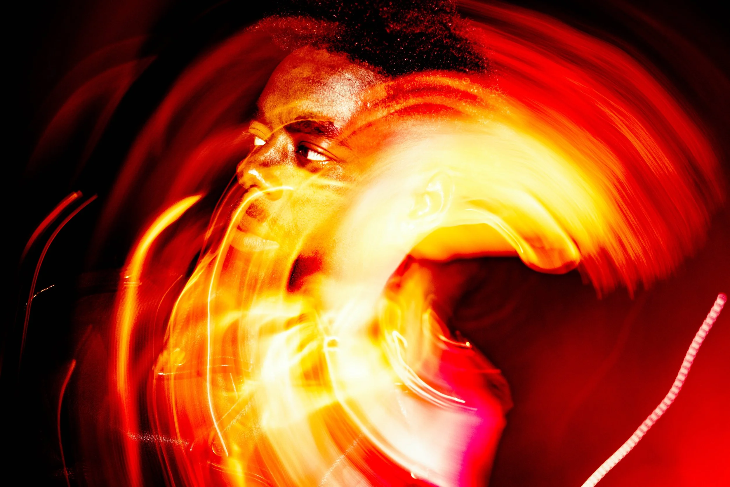 A person with short hair looking to the side in front of swirling bright orange, yellow, and red lights.