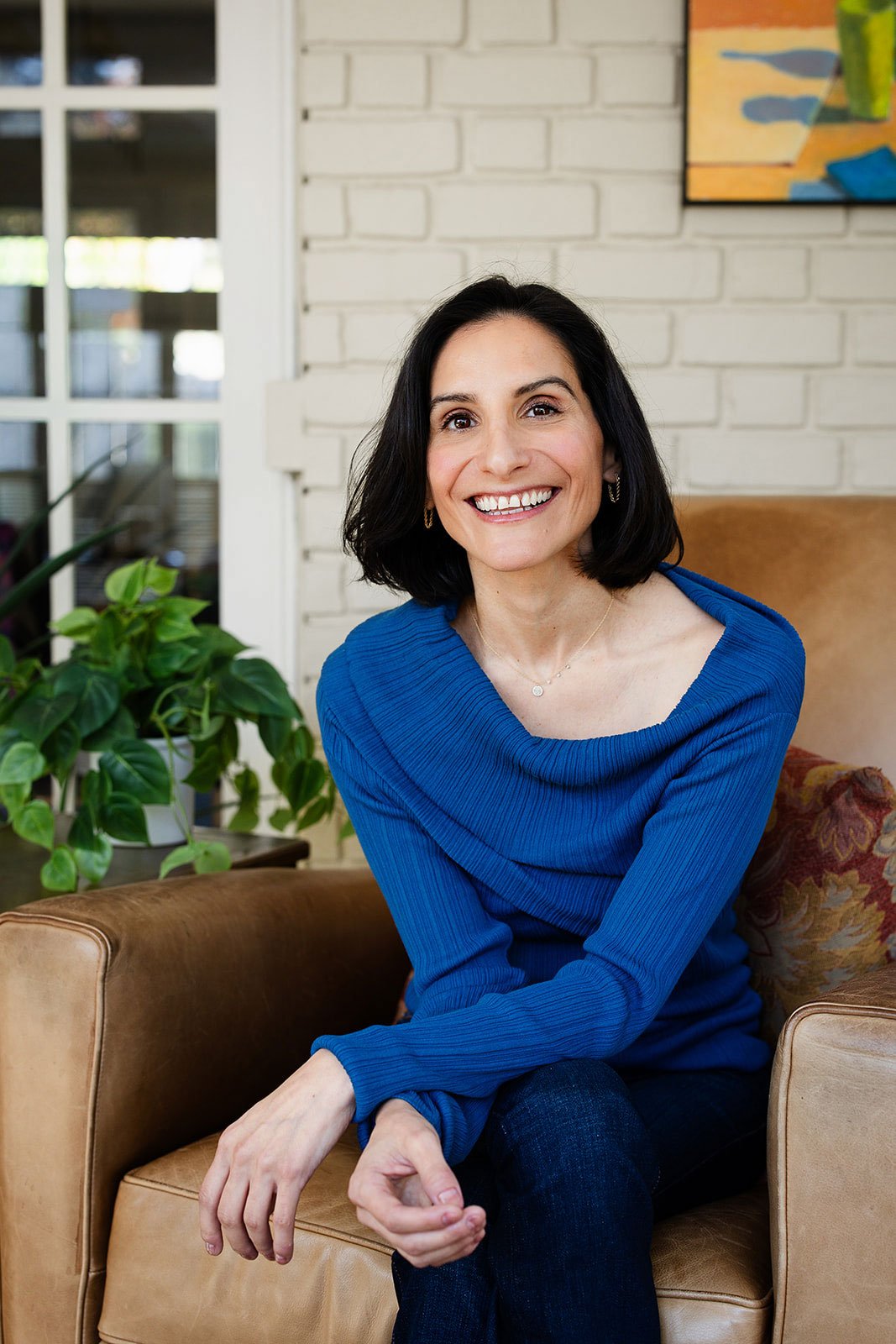Whitney Light, CDFA | Smiling woman in a blue sweater sitting in a brown leather armchair beside a houseplant