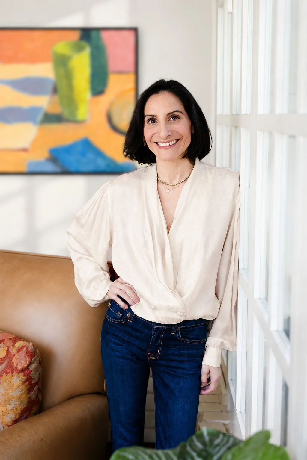 Whitney Light, CDFA | Smiling woman in a cream blouse and dark jeans standing beside a bright window in a modern living room