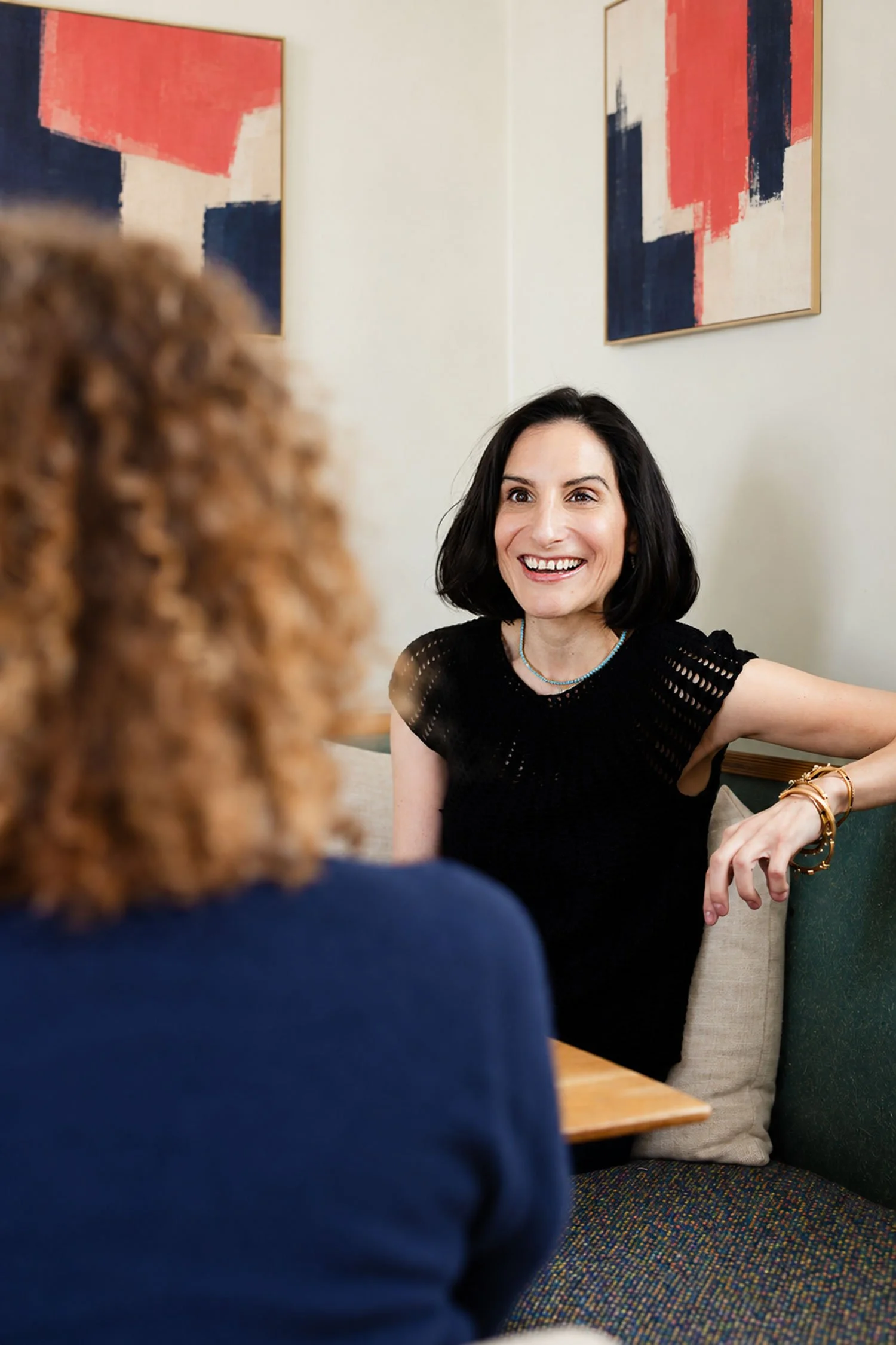 Whitney Light wiht a client | Smiling woman in a black top talking with another person in a cozy seating area decorated with abstract artwork.