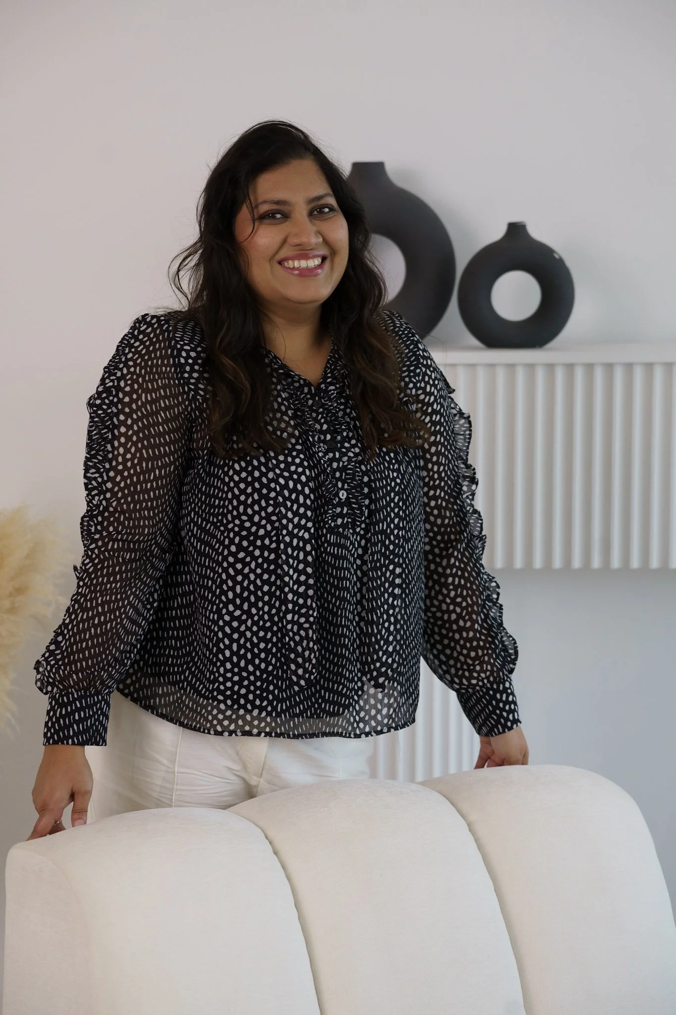 A woman with long wavy dark hair, wearing a black blouse with white polka dots, standing in front of a white sofa with a minimalist decor background that includes two black vases on a shelf and white vertical paneling.