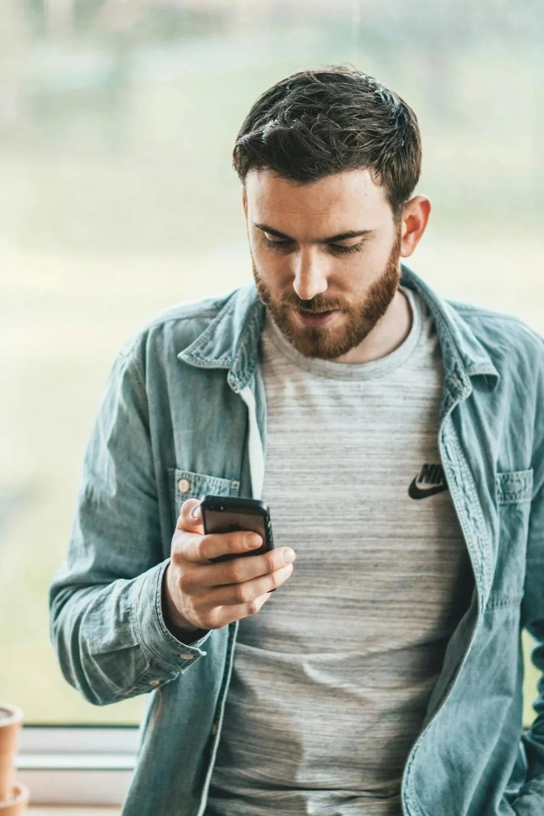 A young man with a beard standing indoors, looking at his phone.