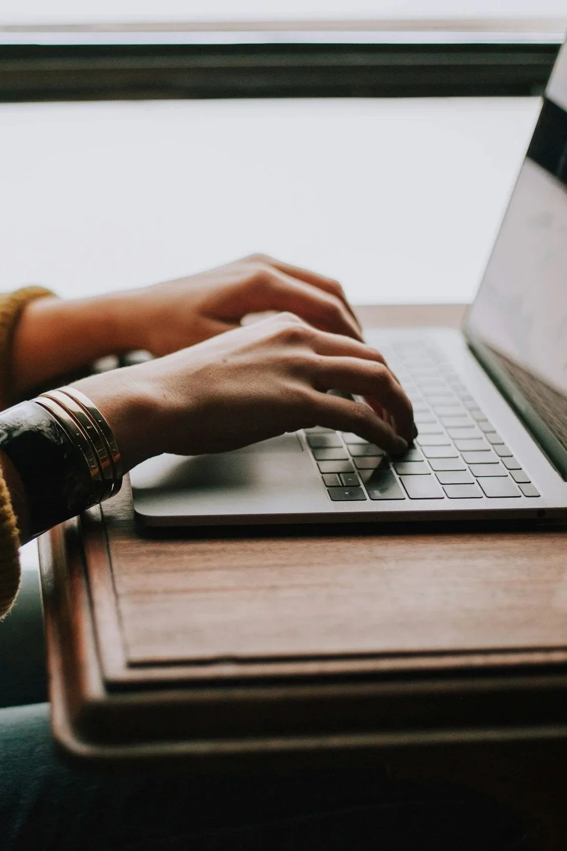 Person typing on a silver laptop keyboard, seated at a wooden desk with a light-colored surface.