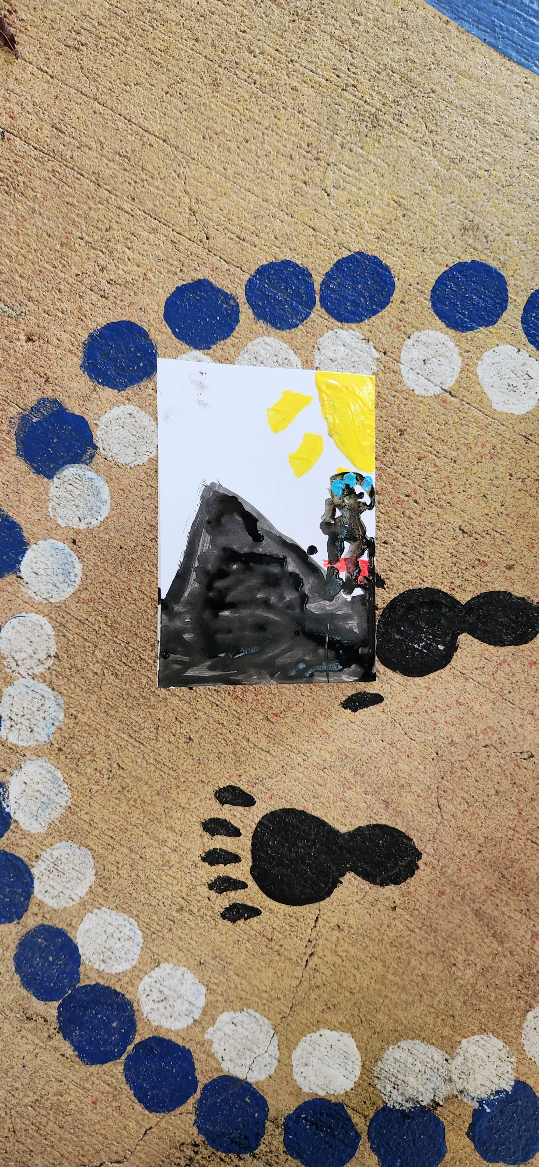 A small painted picture on a beige surface with black, white, blue, and yellow paint. The painting depicts a snow-capped mountain, a yellow sun, blue sky, and a figure near the mountain.