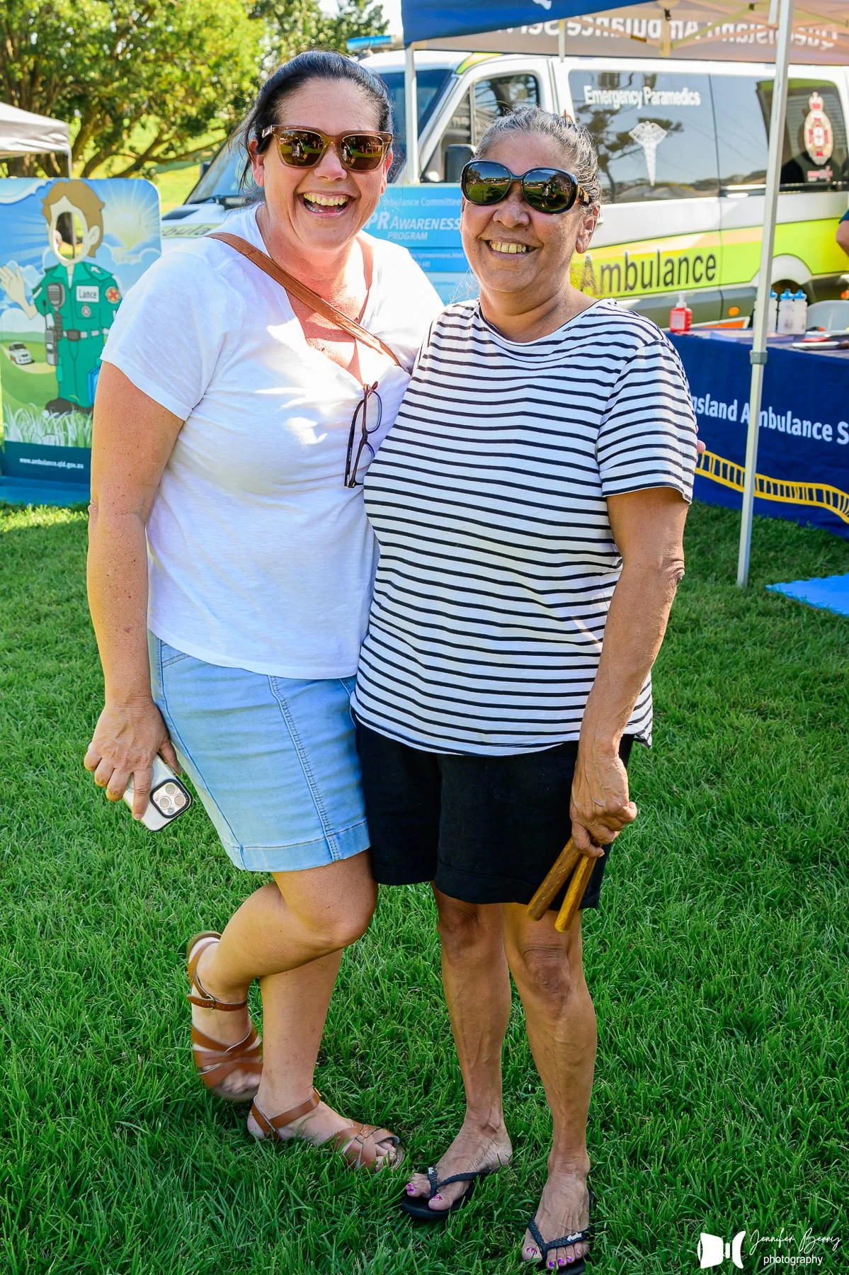 Two women smiling and standing close to each other outdoors on green grass, with emergency response vehicles and posters in the background, under a clear sky.