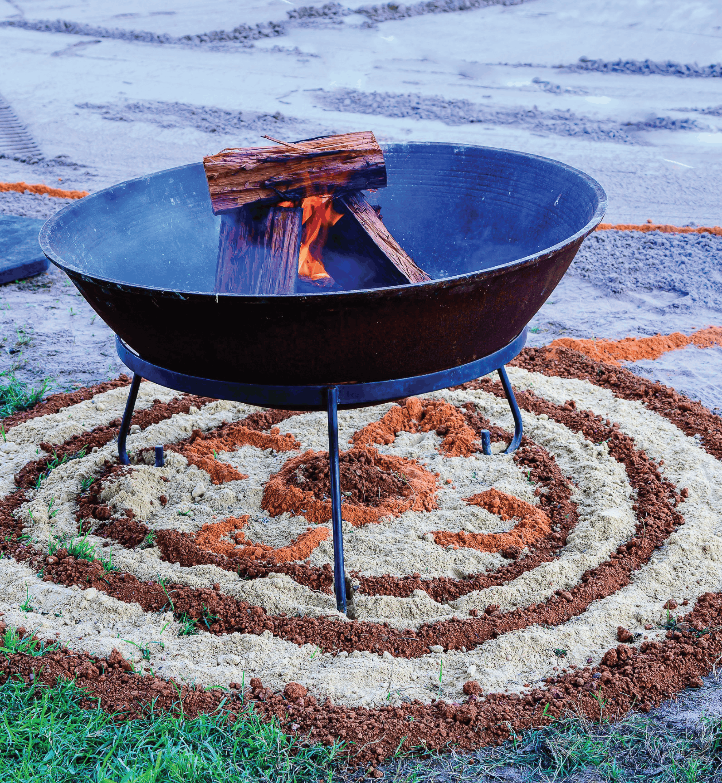 A fire pit with burning wood, surrounded by neatly arranged soil and small green plants, outdoors on a gravel surface.