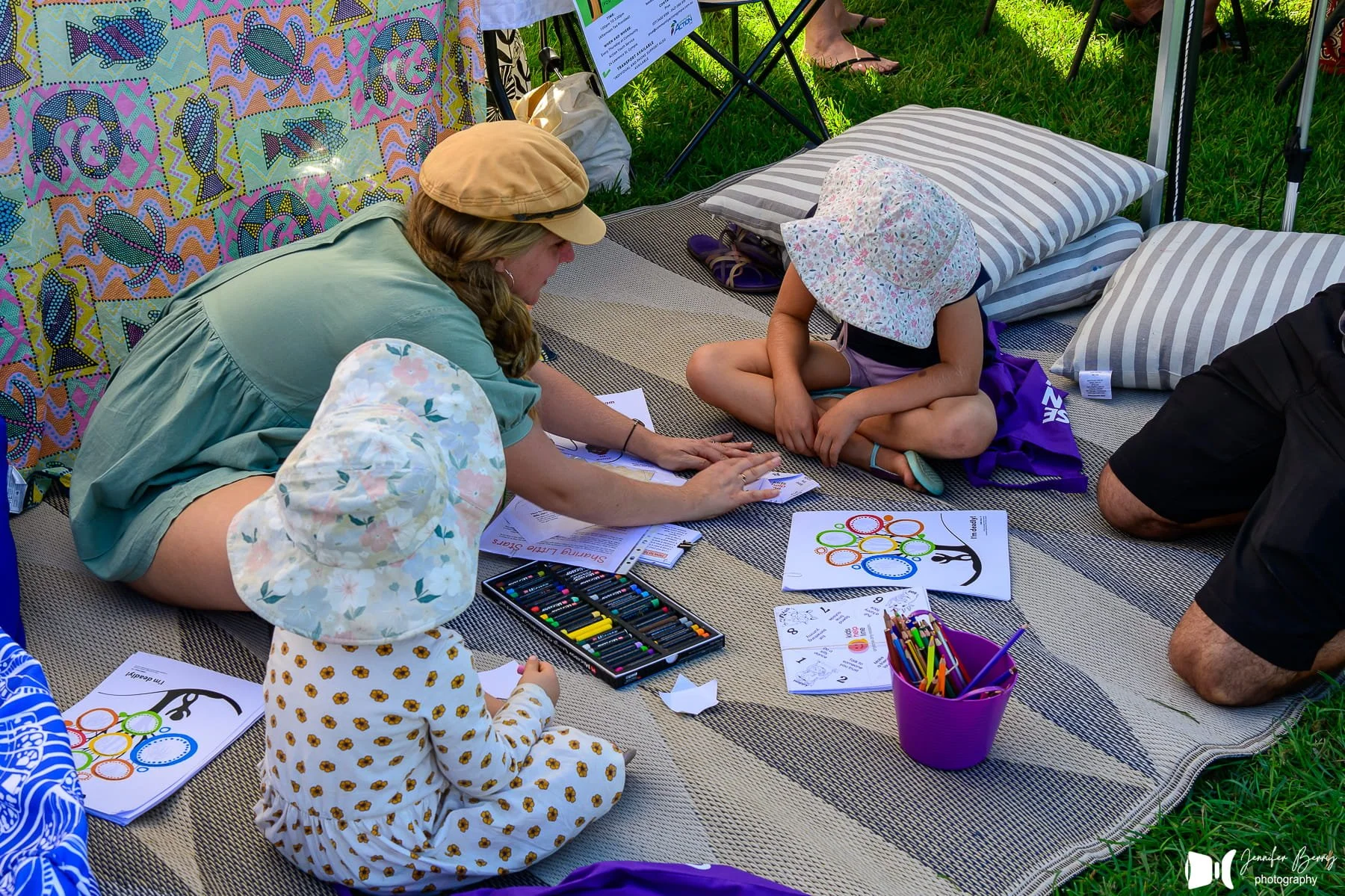 A woman and a young girl are sitting on a blanket outdoors, engaging in arts and crafts activities. The woman is helping the girl with drawing or coloring. There are colored pencils, markers, and worksheets around them. The girl is wearing a floral h
