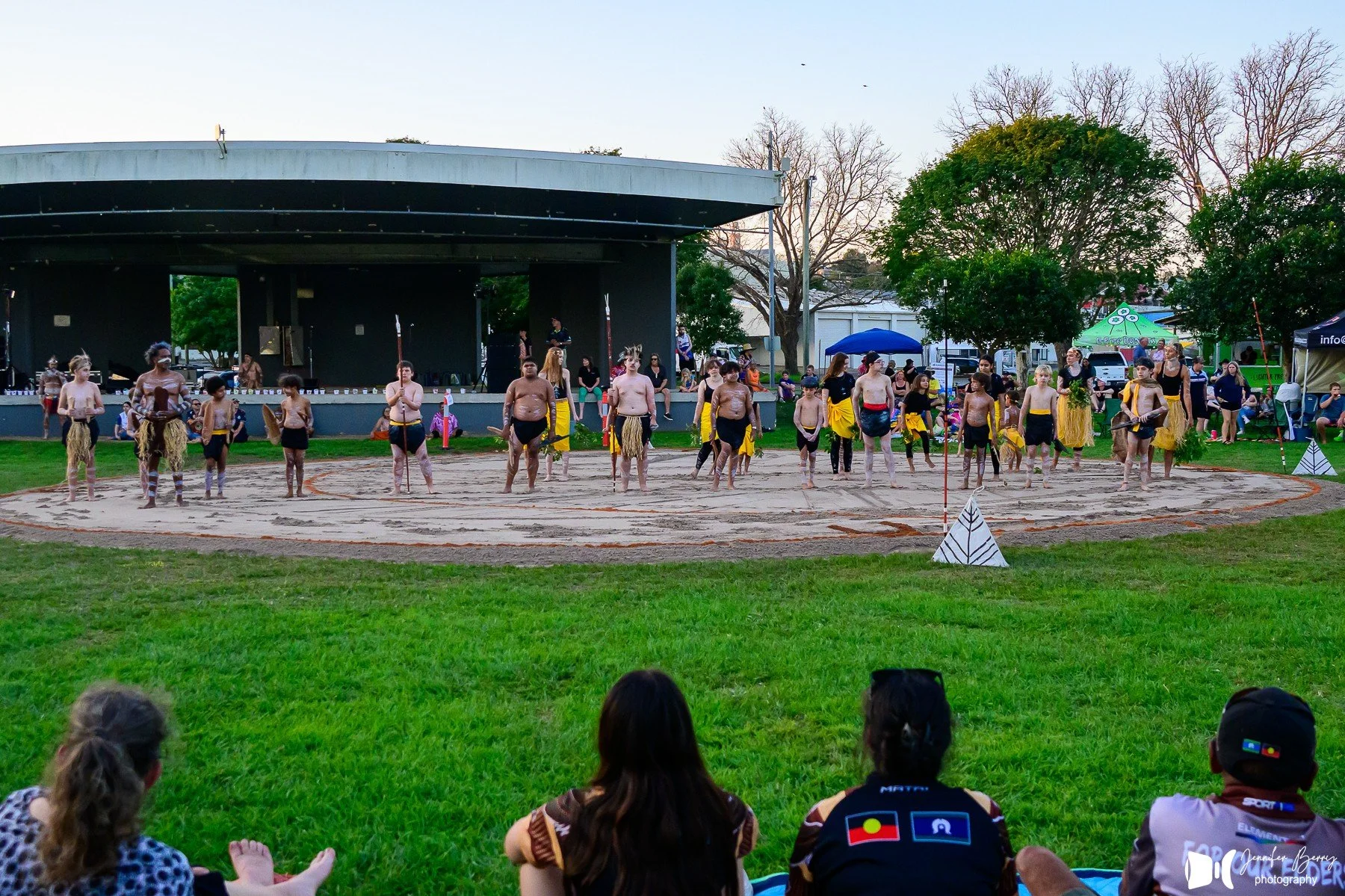A group of children dressed in traditional Pacific Islander costumes performing a dance on a sand circle in a park, with spectators watching and tents in the background.