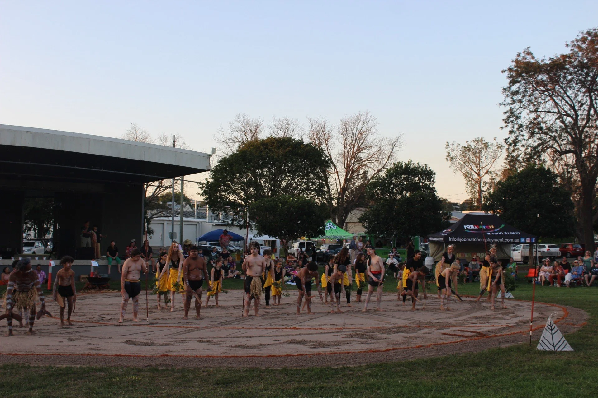 People performing a cultural dance in an outdoor park during daytime with tents and trees in the background.