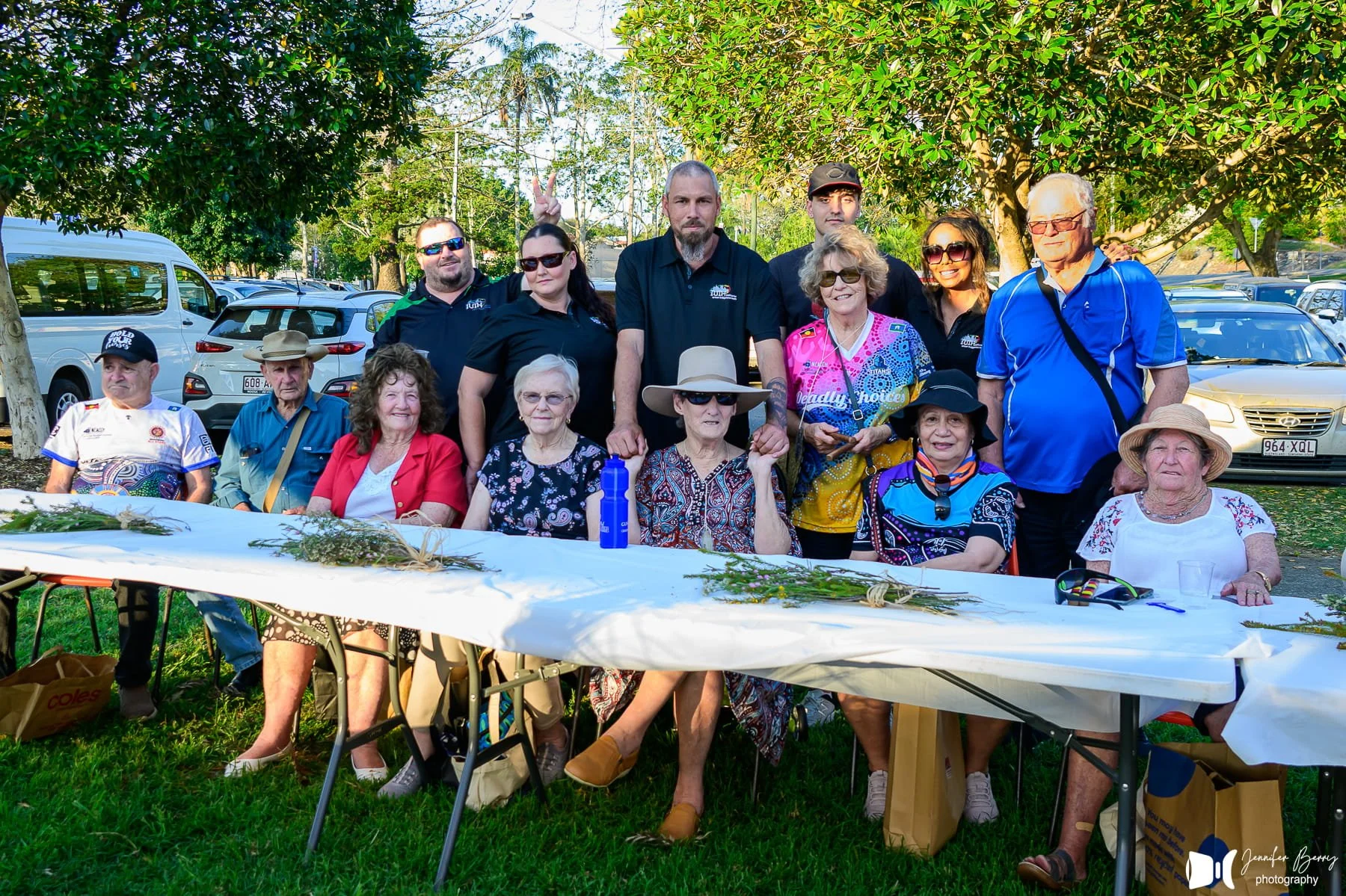 A group of seniors and younger adults gathered around a long table outdoors, celebrating an event in a park with cars parked in the background and trees.