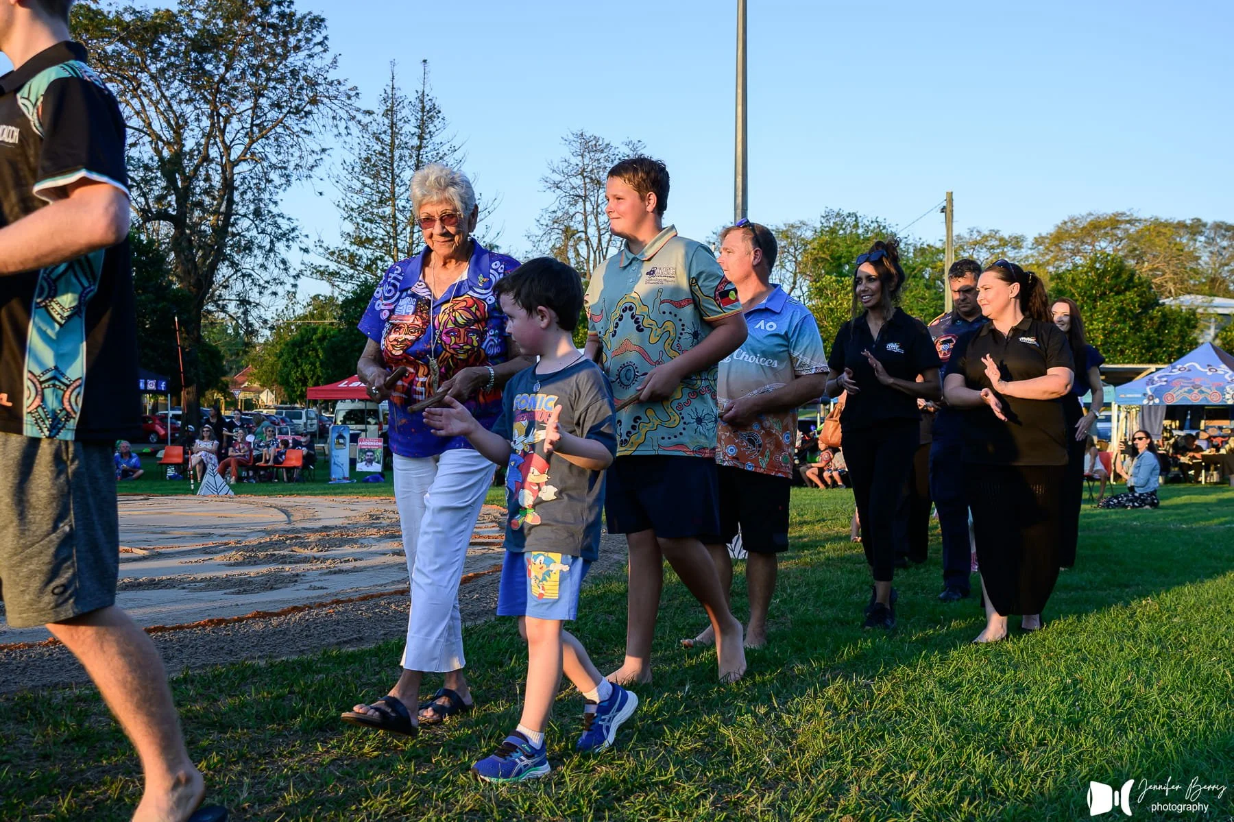 People walking in a park during an event, with some sitting on benches and others clapping, under a clear sky with trees in the background.