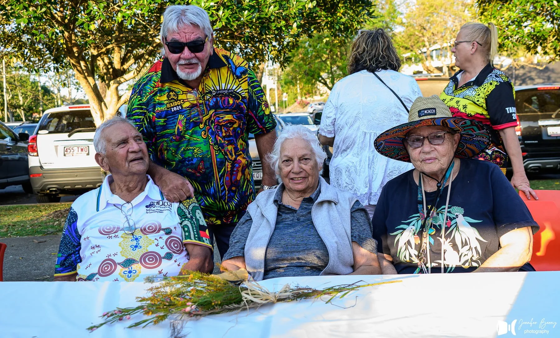 Group of five elderly people seated outdoors with a table in front, outdoors, with trees and parked cars in the background. One woman is wearing a large colorful hat, and another woman is wearing sunglasses and a patterned shirt. Two women are standi