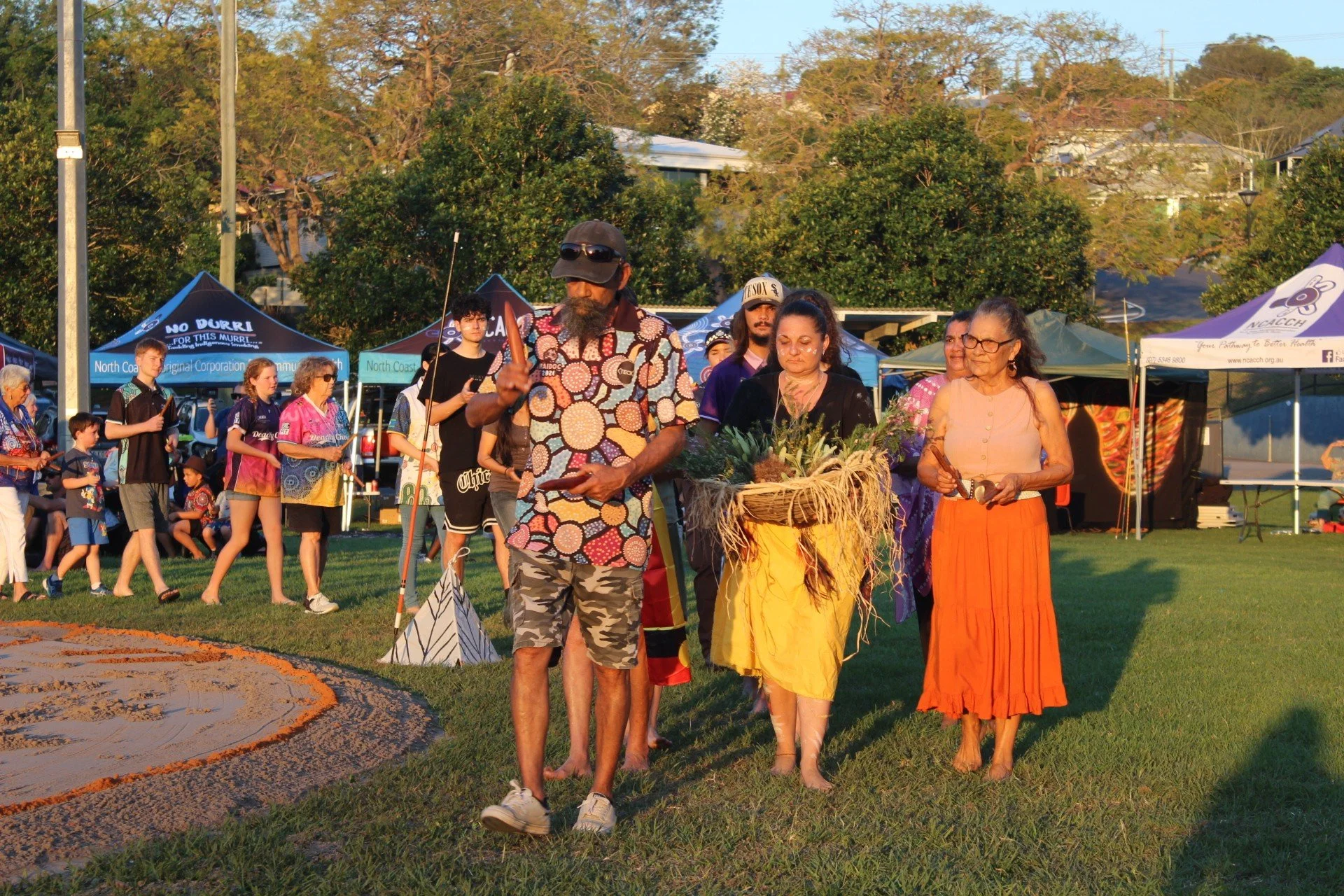 A group of Indigenous people participating in a cultural ceremony outdoors with tents in the background.