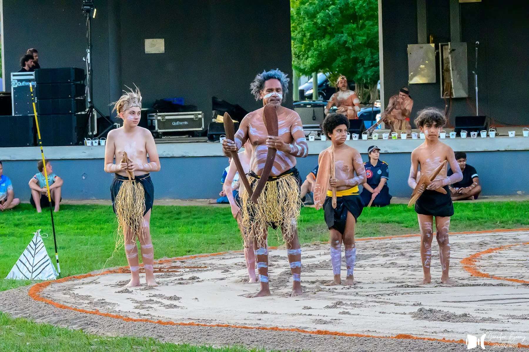 A group of children and an adult dressed in traditional Indigenous clothing, standing in a semi-circle on a patterned ground, possibly for a cultural performance or presentation.