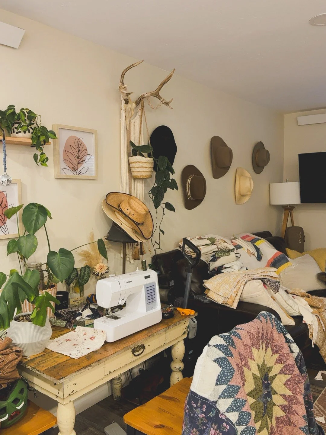 Cluttered living room with a table holding a sewing machine, plants, and hats on the wall and sofa.