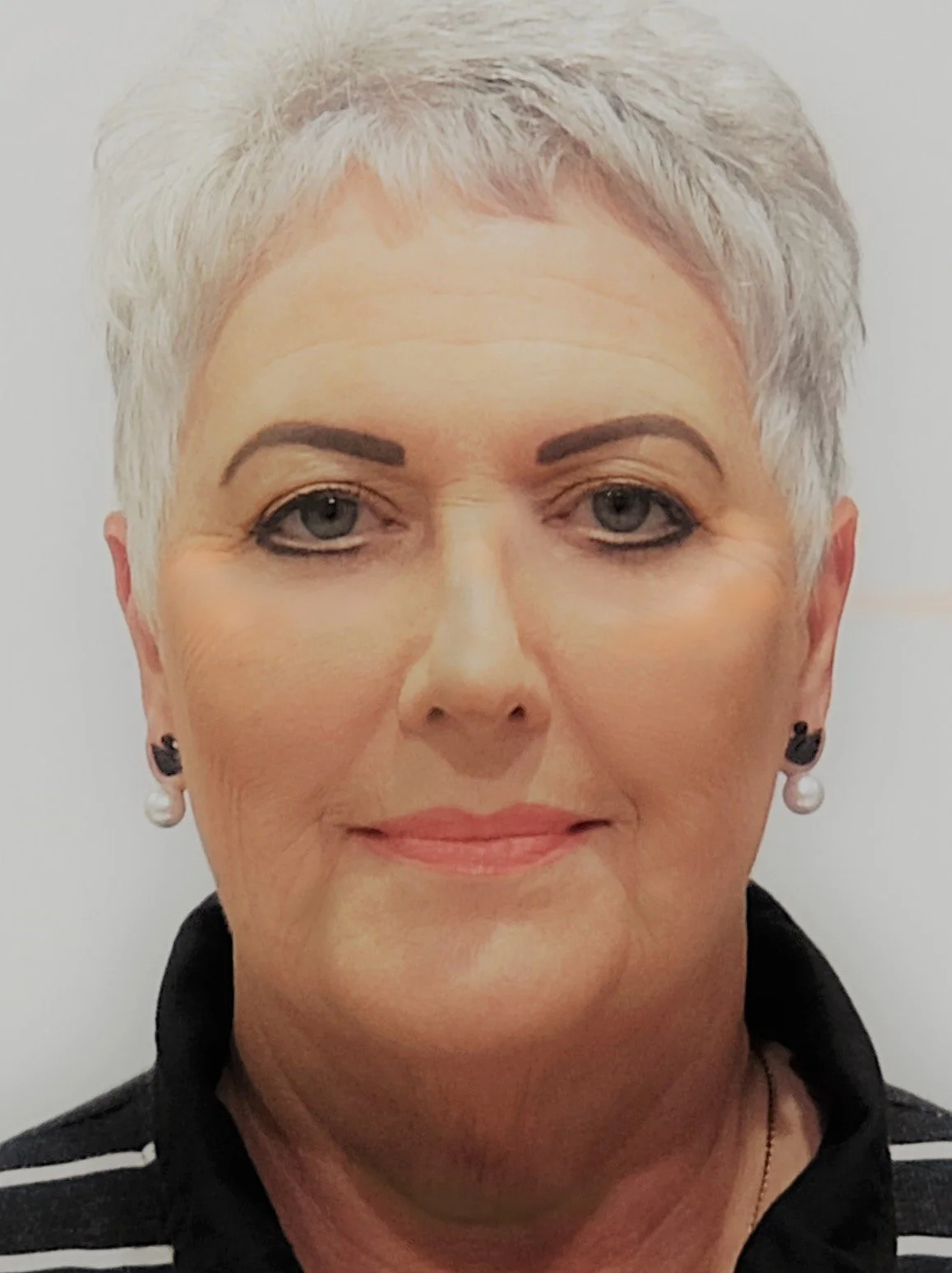 Close-up of a mature woman with short, gray hair and pearl earrings, wearing a black and white striped top, against a plain background.