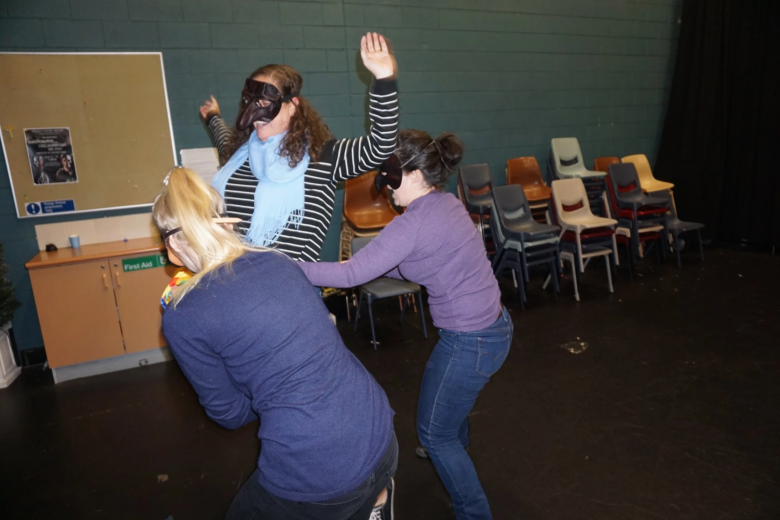 Three women wearing masks pose playfully in a community hall with stacked chairs.